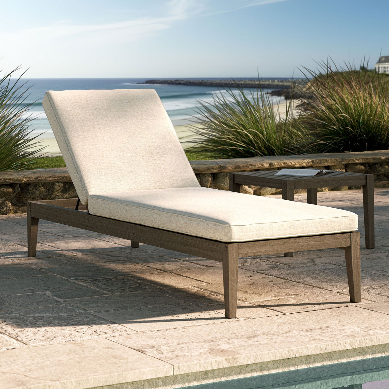 Outdoor teak chaise lounge with light pebble cushion styled by a pool with a serene coastal backdrop || EEI-7256-WGR-PEB || front_angle_converted_room