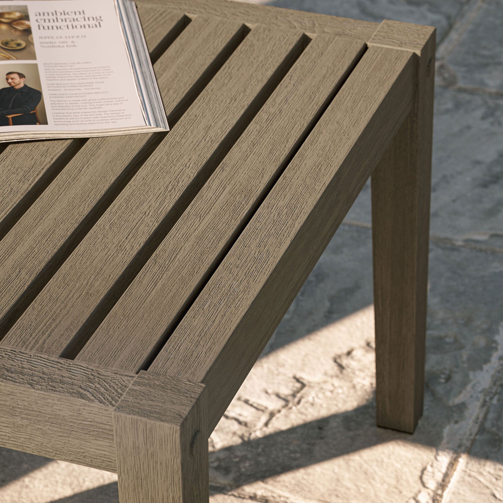 Teak side table styled on a sunlit stone patio, with slatted top holding an open magazine for relaxed lounging. || EEI-7258-WGR || detail_room