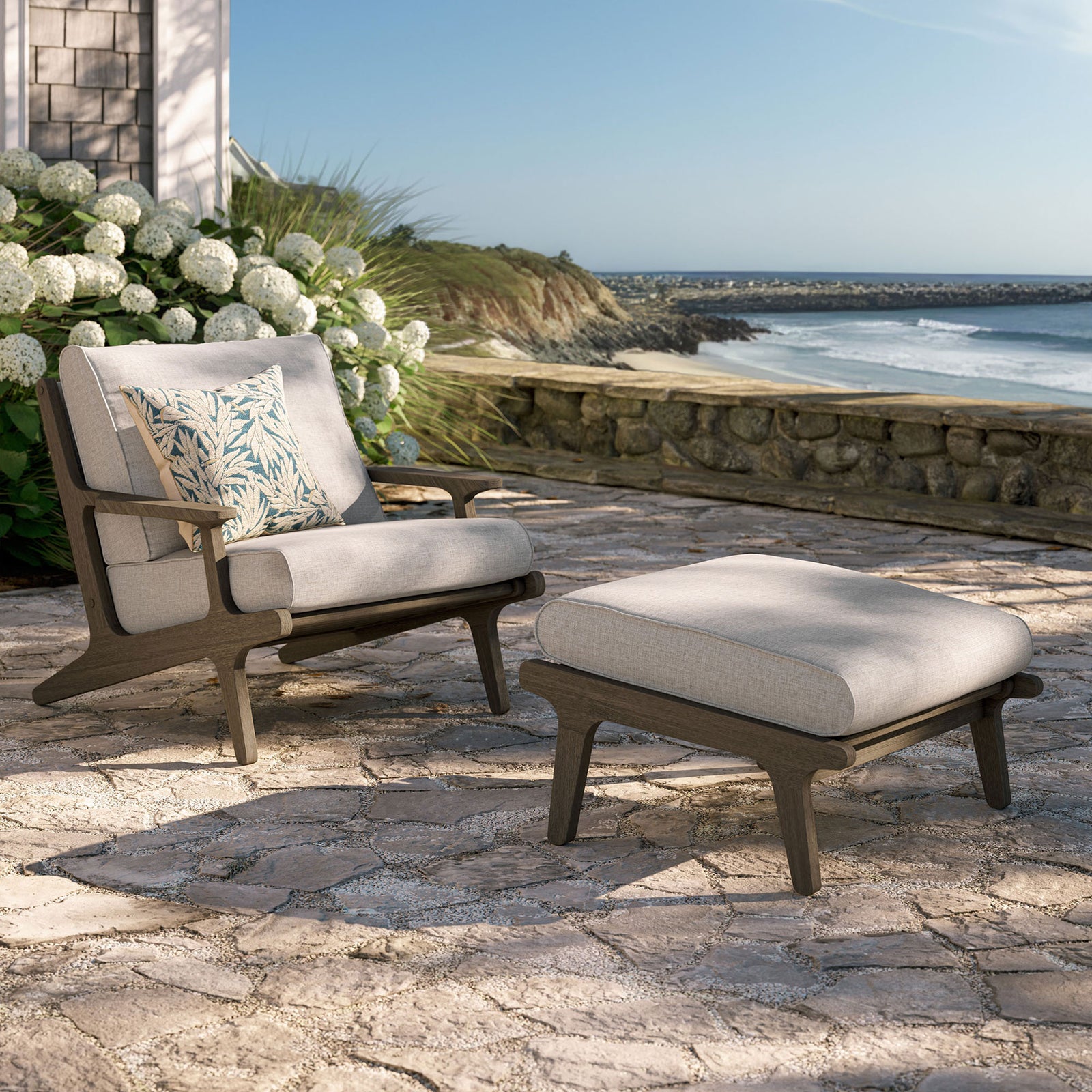 Saratoga pebble chair and ottoman styled on a sunny patio with ocean views and lush hydrangeas || EEI-7268-WGR-PEB || front_angle_room