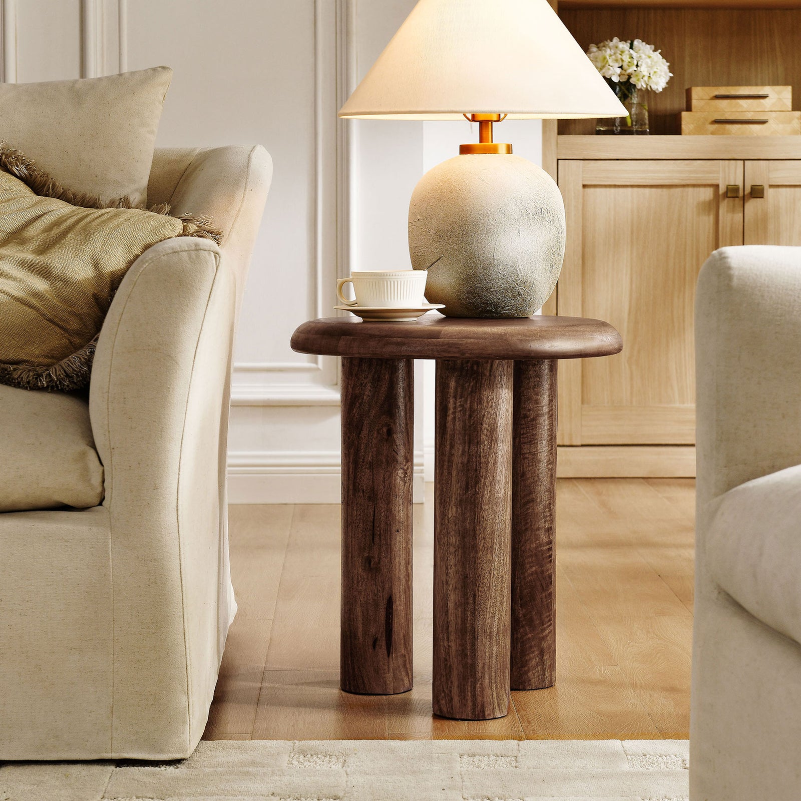 Round mango wood side table in a bright living room, perfect for holding a lamp and everyday essentials. || EEI-7284-WAL || front_angle_room