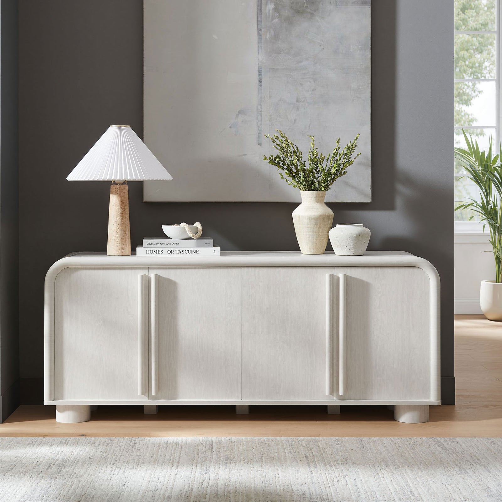 Curved ash sideboard styled in a contemporary living room with lamp, books, and ceramic vases || EEI-7315-ASH || front_room
