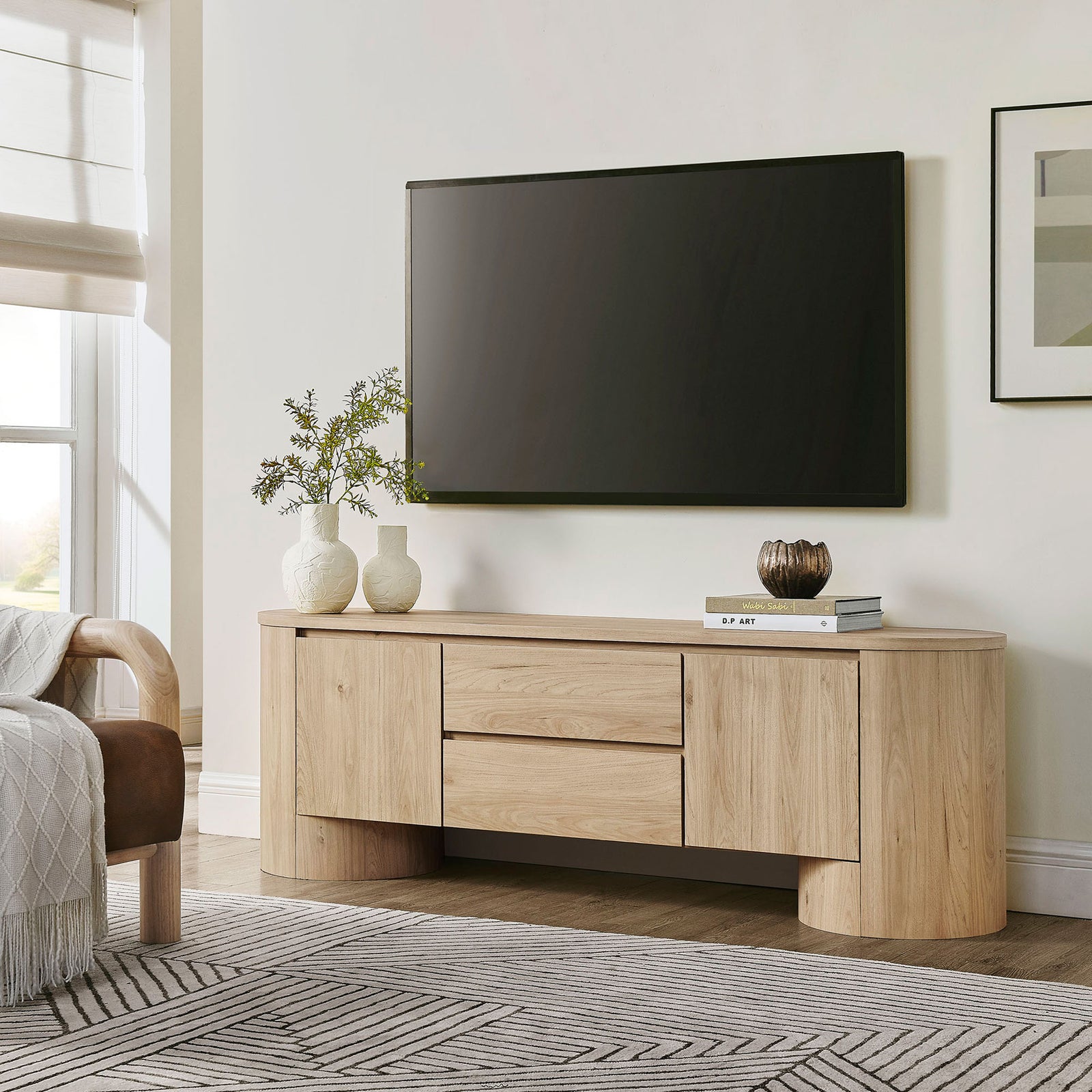 Light oak oval TV cabinet positioned under a wall-mounted screen in a bright contemporary living room || EEI-7440-LOA || front_angle_room