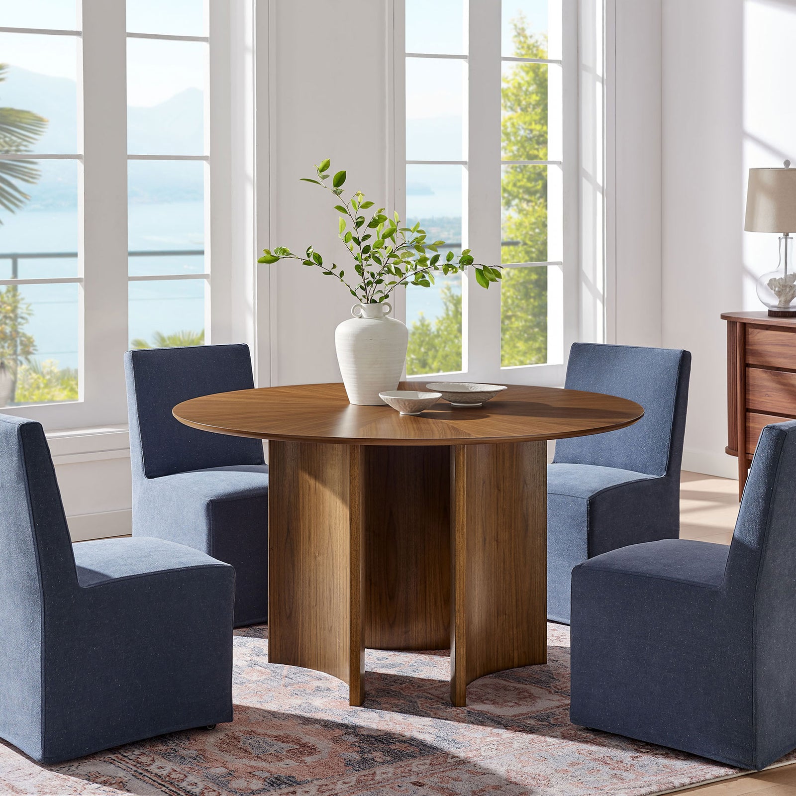 Walnut round dining table with curved base surrounded by four blue chairs in an airy dining room || EEI-7603-WAL || front_angle_room