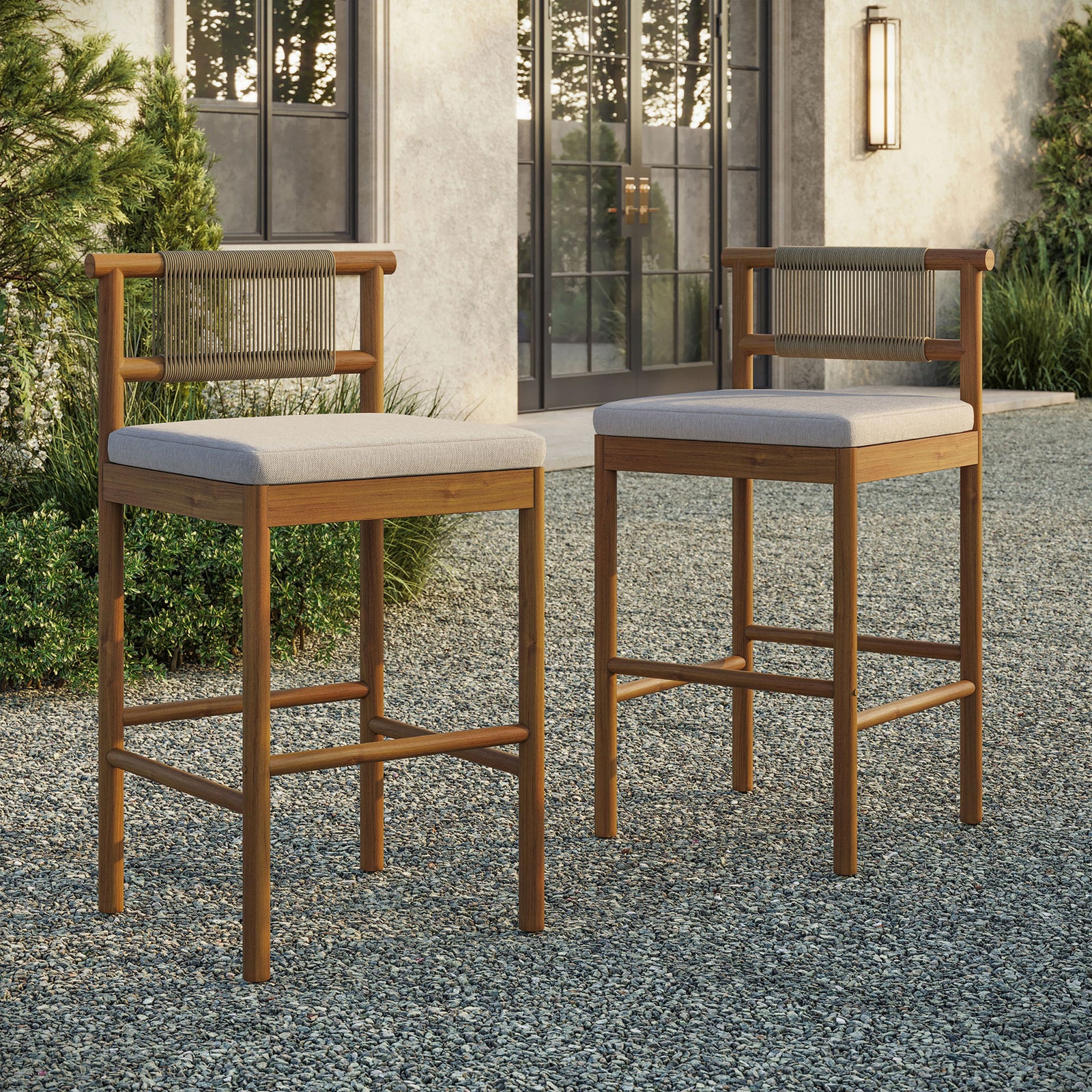 Pair of teak bar stools grouped around an outdoor bar table on a gravel terrace || EEI-7626-NAT-SAI || front_angle_multi_room
