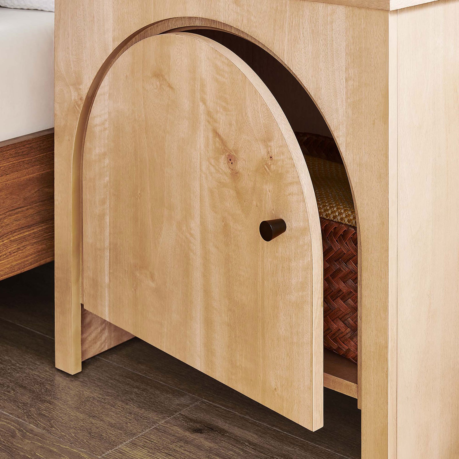 Arched oak nightstand beside a bed with door open to show woven basket storage inside. || MOD-7109-OAK || front_angle_open_room