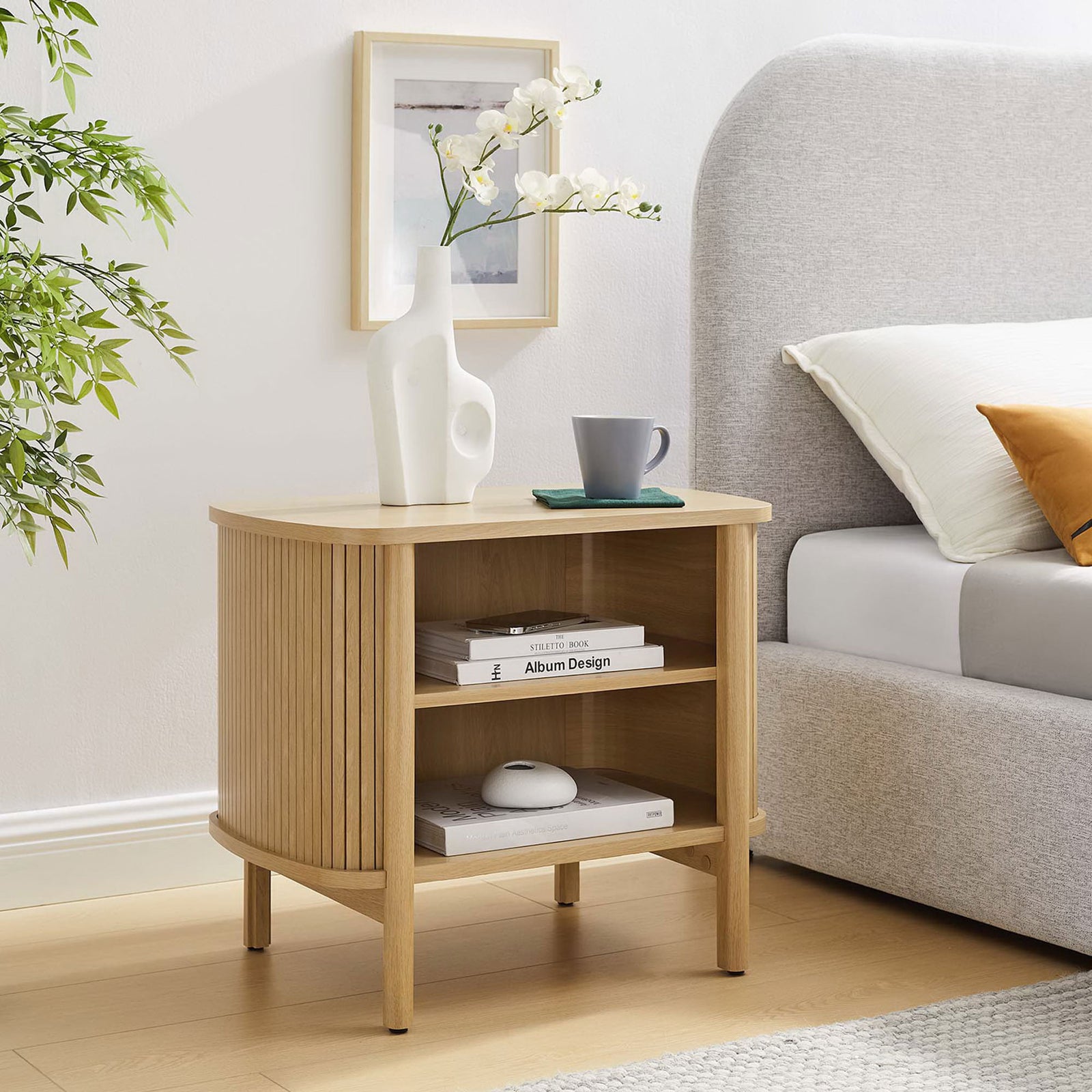 Cadence oak nightstand beside a modern upholstered bed, styled with decor to showcase its two open storage shelves || MOD-7157-OAK || front_angle_room