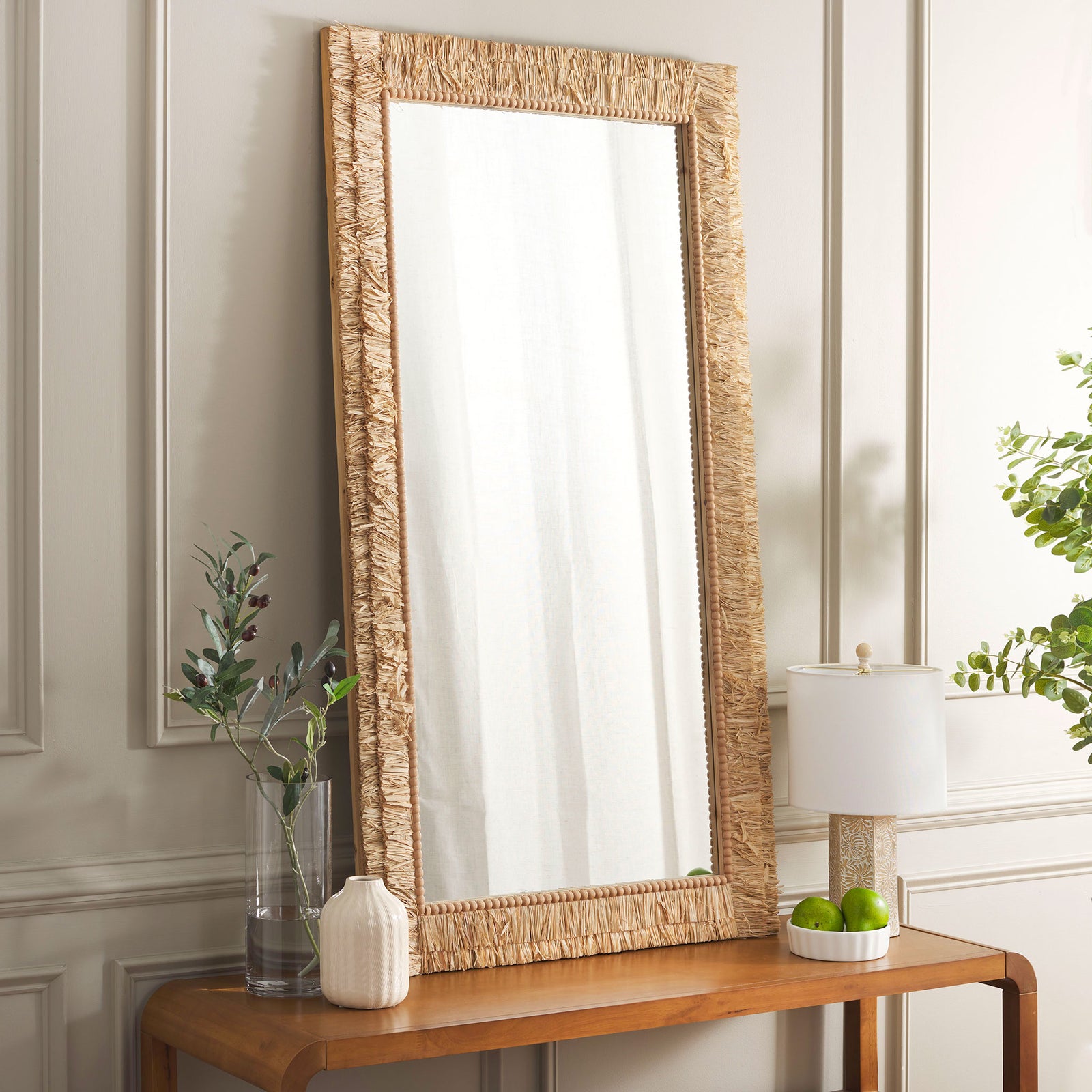 Warm natural texture frames the tall mirror resting on a wood console in a neutral room. || MRR9003A || front_angle_room