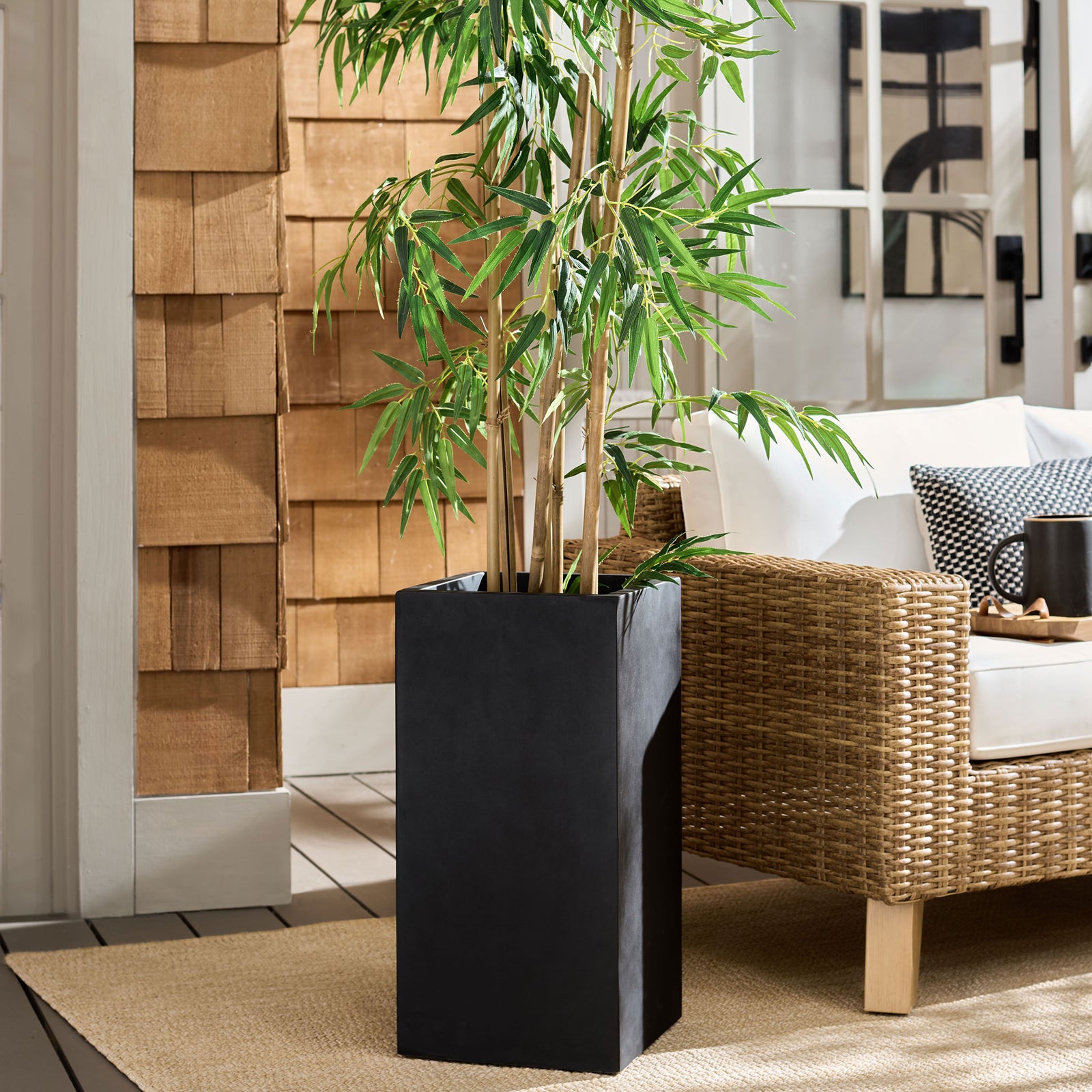 Tall black planter adds a sleek contrast to leafy greenery and woven patio furniture || PAT1402A || front_angle_room