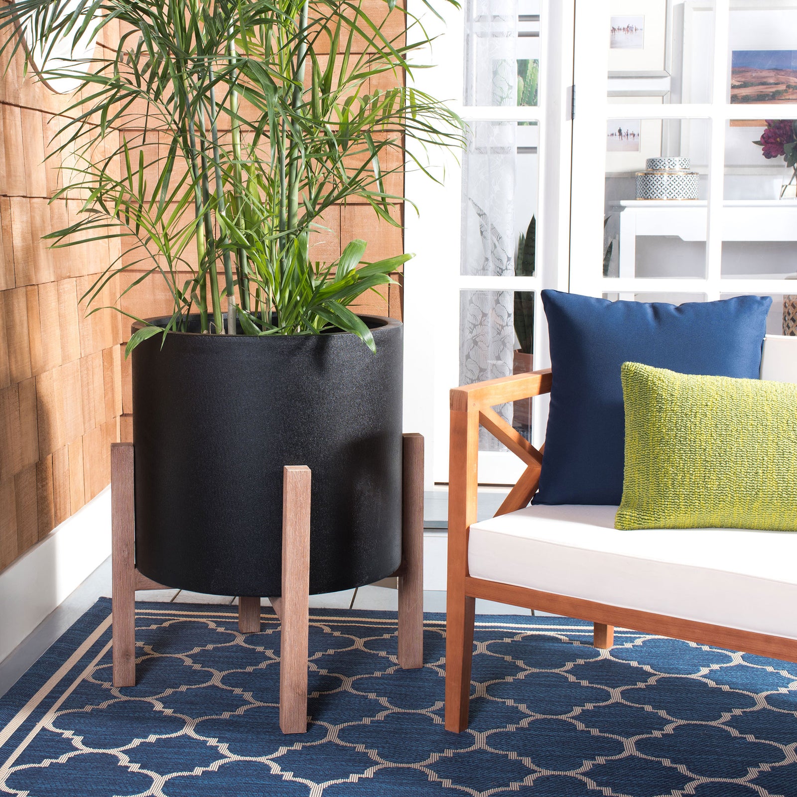 Tall greenery rises from a generous black planter on a wood-toned stand beside a lounge chair. || PAT1515A || front_room