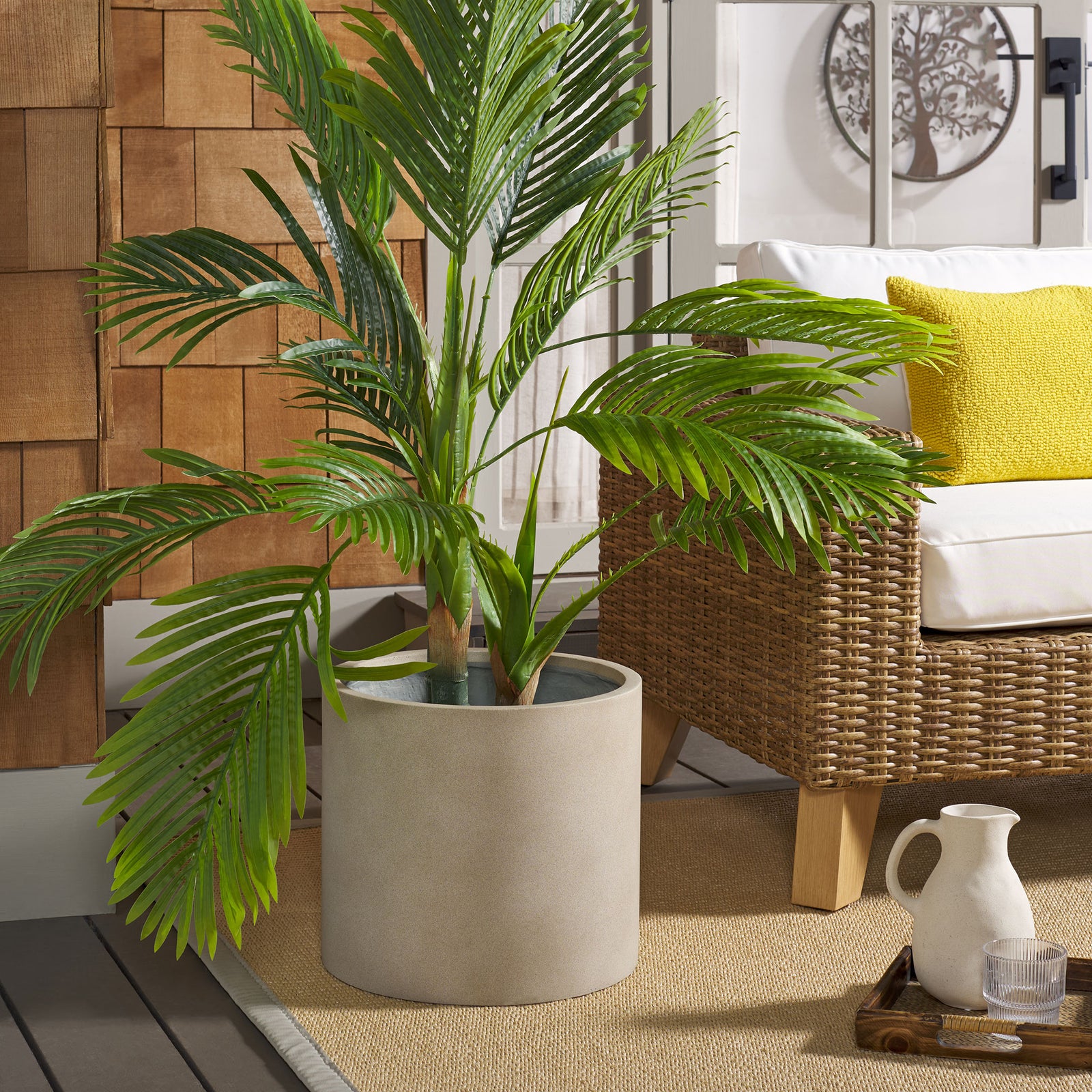 A beige cylindrical planter grounds a leafy palm beside woven patio seating || PAT1516B || front_angle_room