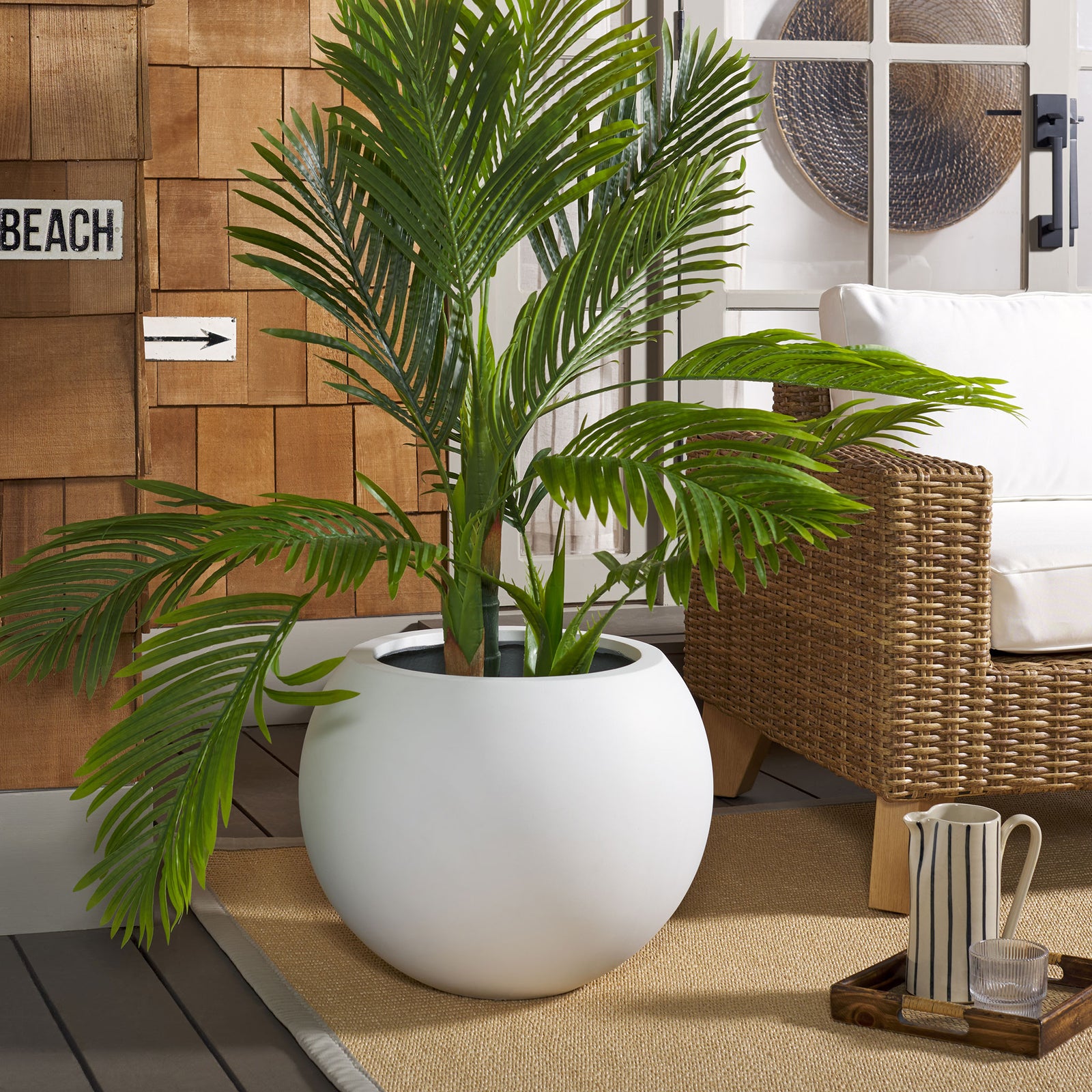 A broad white planter anchors tropical greenery beside wicker seating and a woven rug. || PAT1519C || front_room