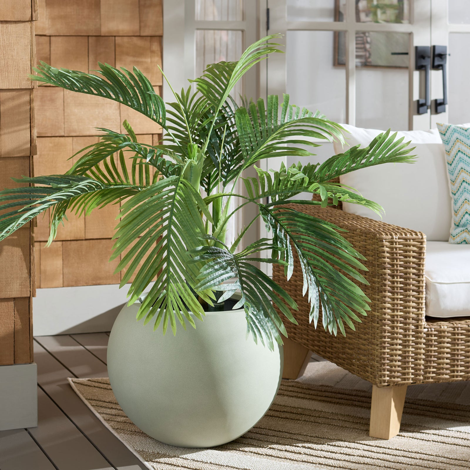 A large sage planter gives a lush palm a sculptural base beside a woven lounge chair. || PAT1519E || front_room
