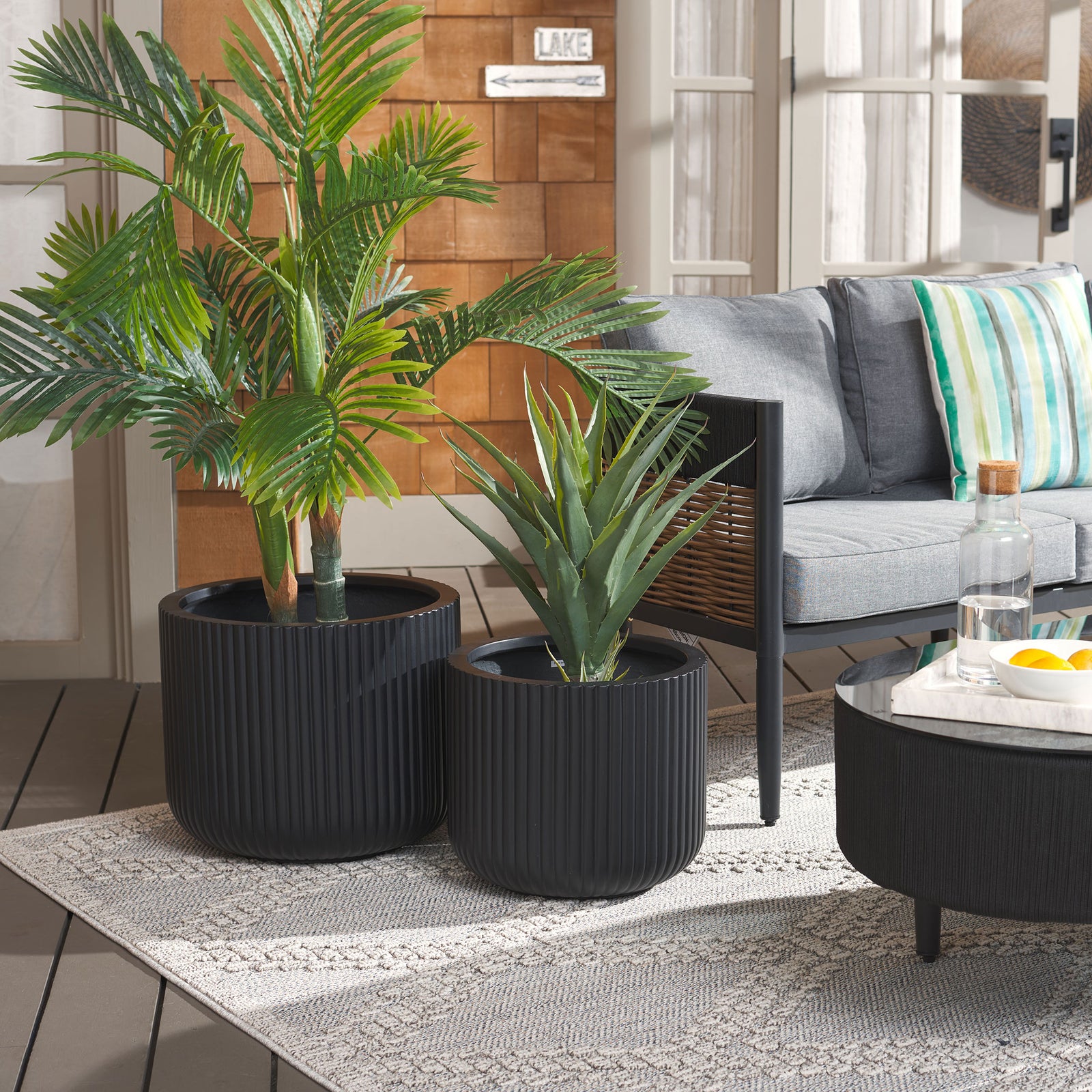Black fluted planters frame a leafy palm and spiky greenery beside a cushioned patio chair. || PAT1532A-SET2 || front_angle_room