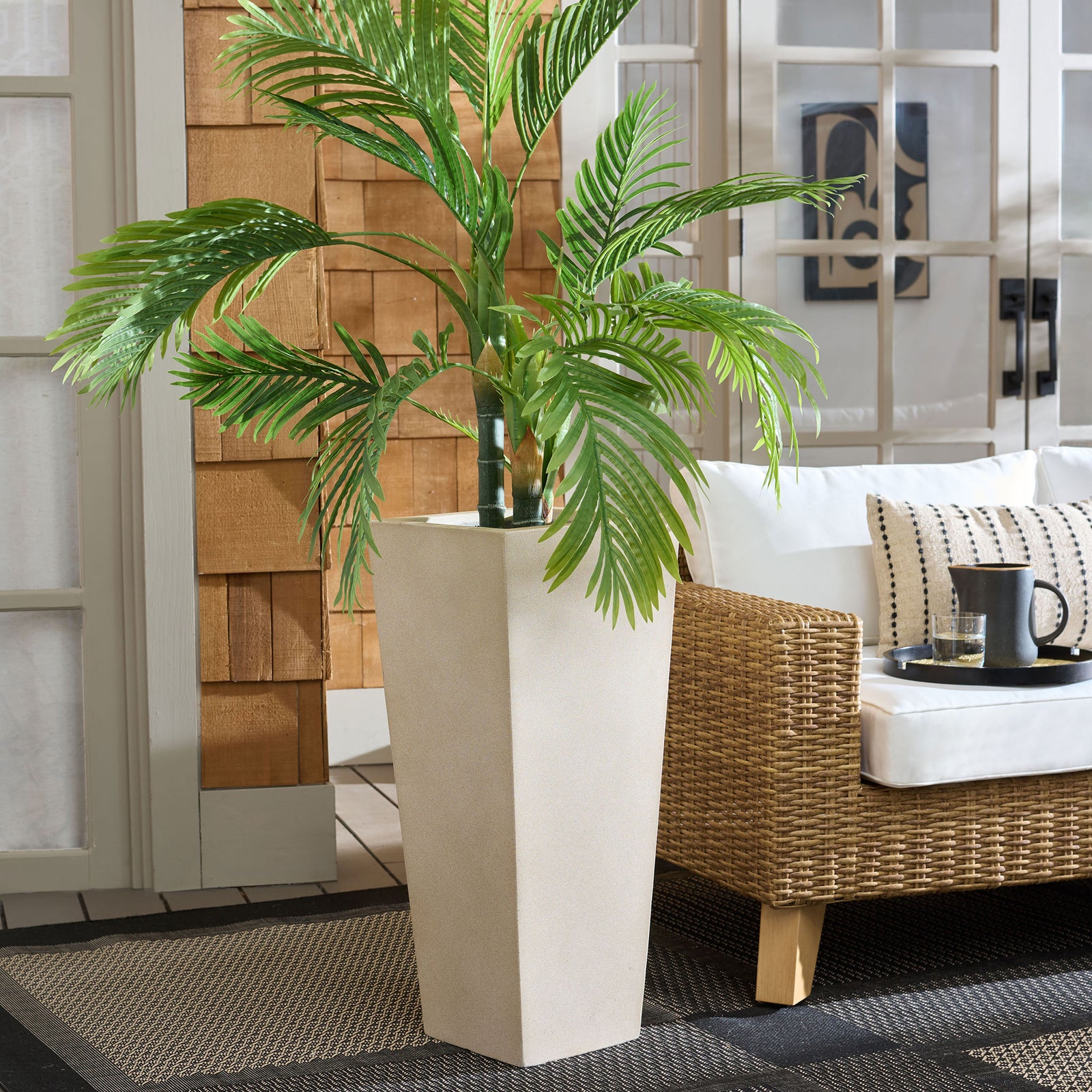 Tropical foliage rises from a tall beige planter beside woven patio seating. || PAT1543B || front_angle_room