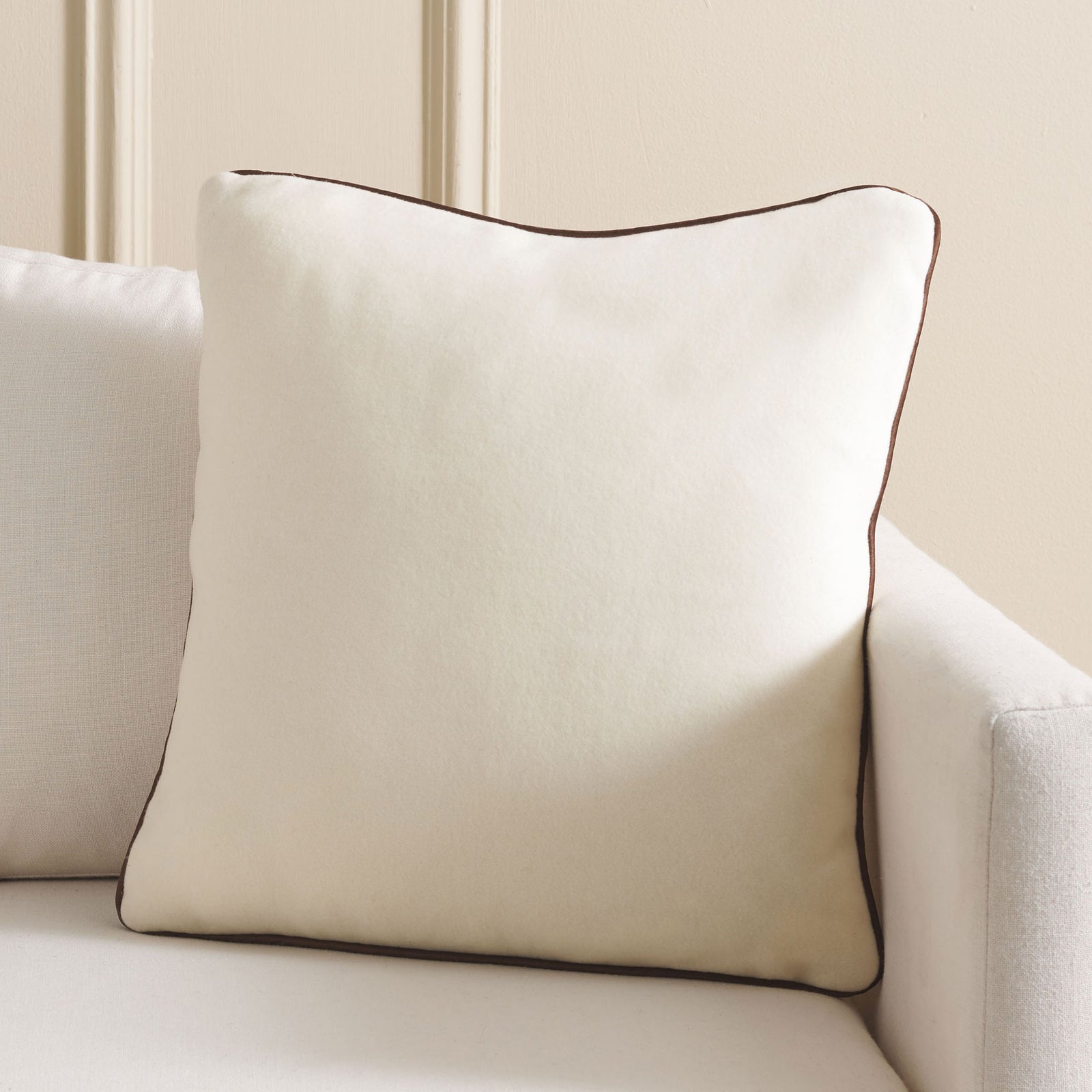 An ivory wool pillow edged in brown piping adds a cozy, tailored accent to a neutral sofa. || PLS7223A-1818 || front_room