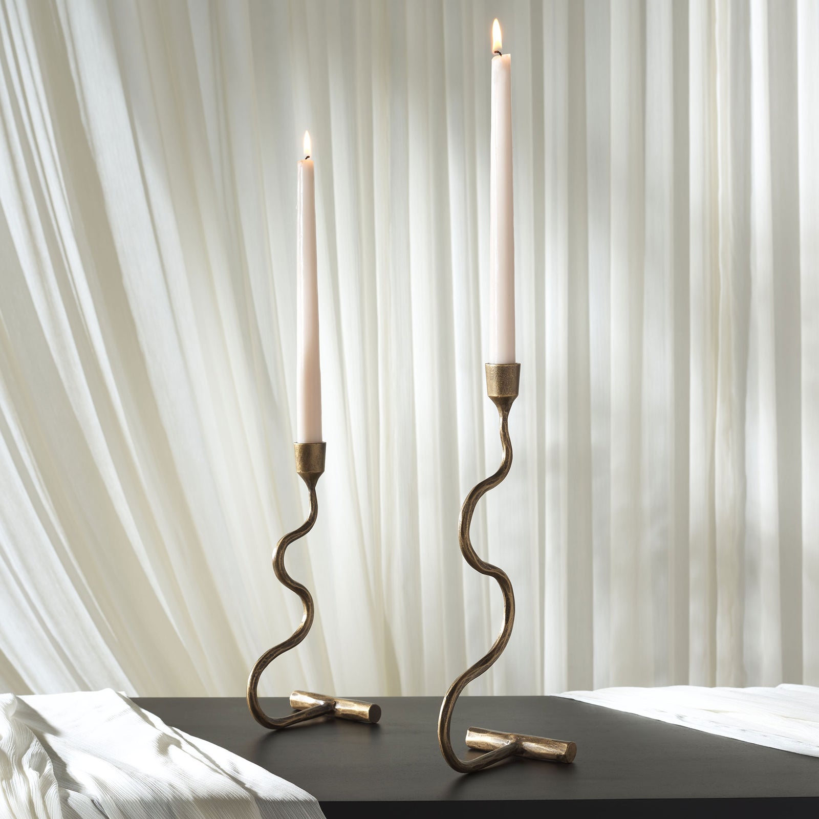 Lit white taper candles lend a soft glow to two sculptural gold metal candlesticks. || RDC2500A-SET2 || front_angle_room