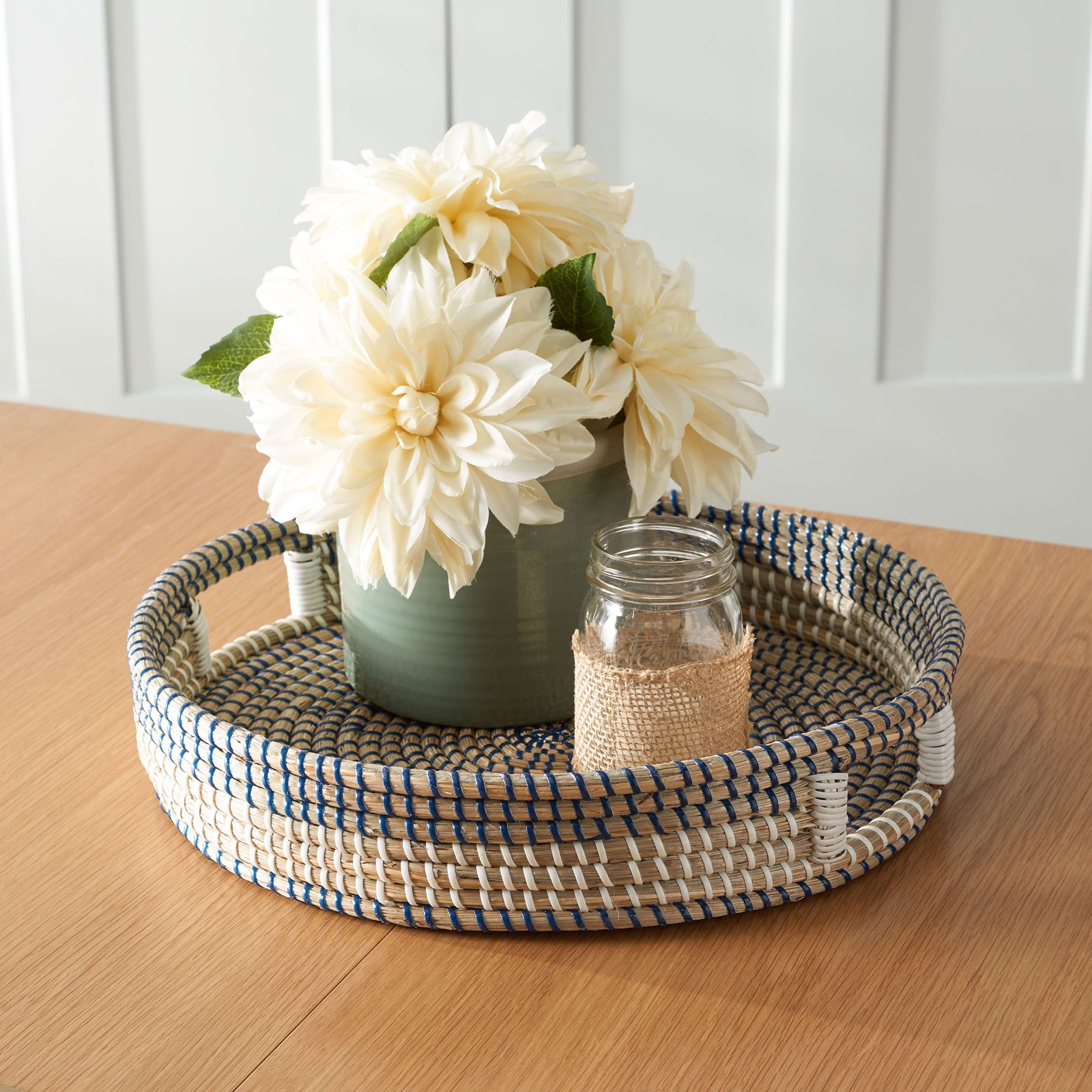 A blue-and-white woven tray holds a flower-filled vase and a small glass jar on a wood table. || STG1809A || front_angle_room