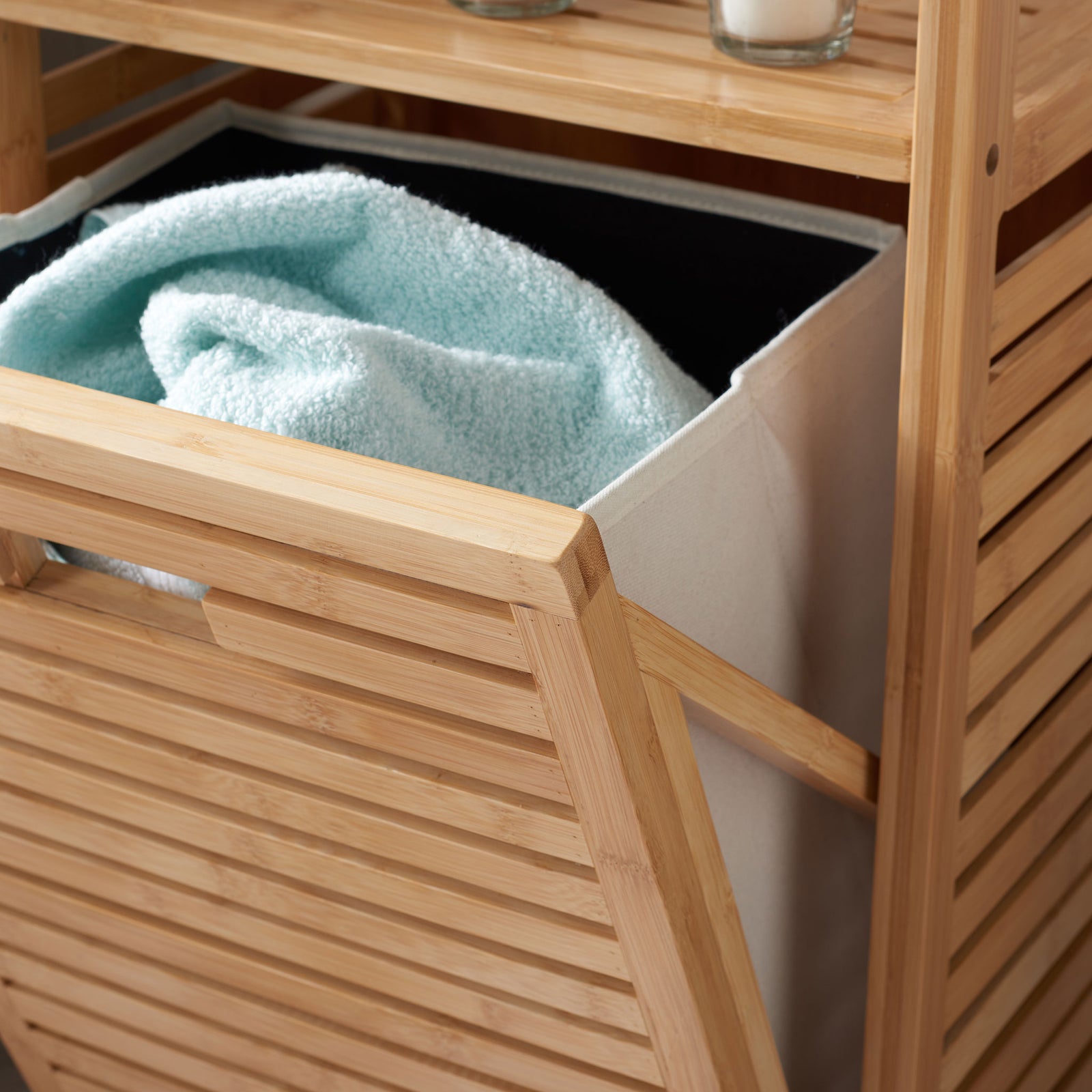 A cream fabric basket with a dark interior cradles towels behind the tilted bamboo hamper front || STG1903A || storage_open_room