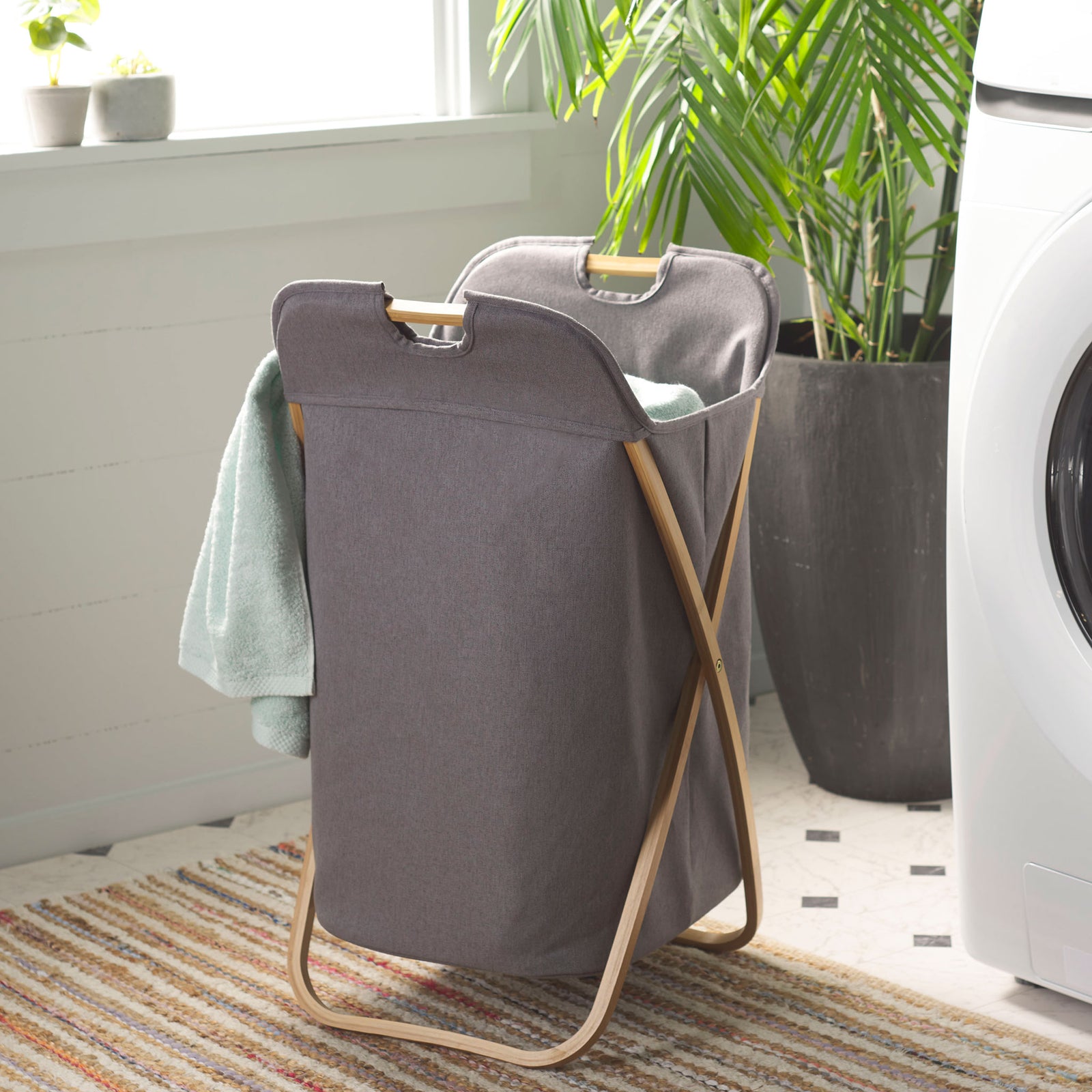 The gray fabric hamper holds laundry beside the washer on its natural bamboo frame. || STG1906A || front_angle_room