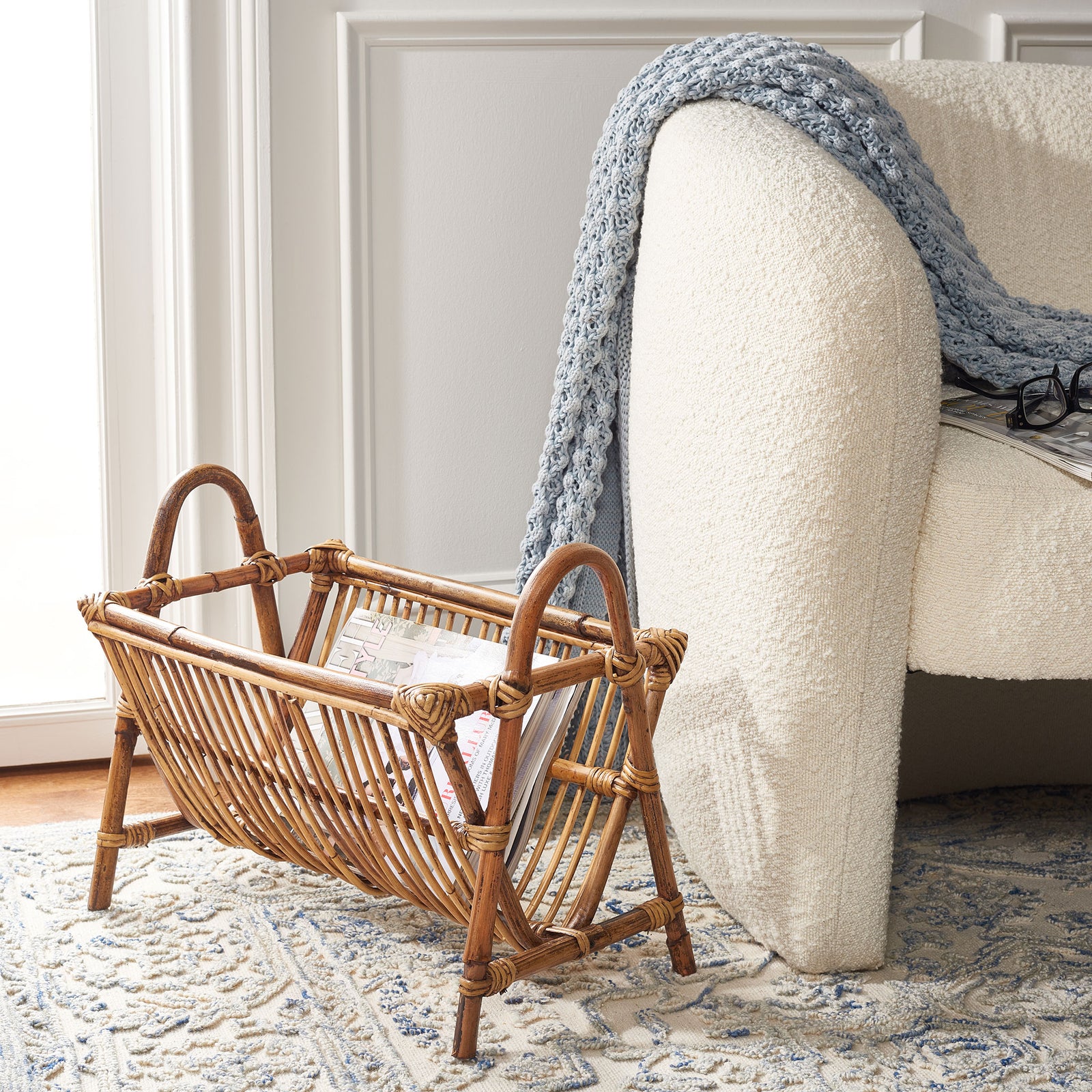 A honey-brown rattan newspaper rack holds reading material beside an upholstered chair || STG6500A || front_angle_room