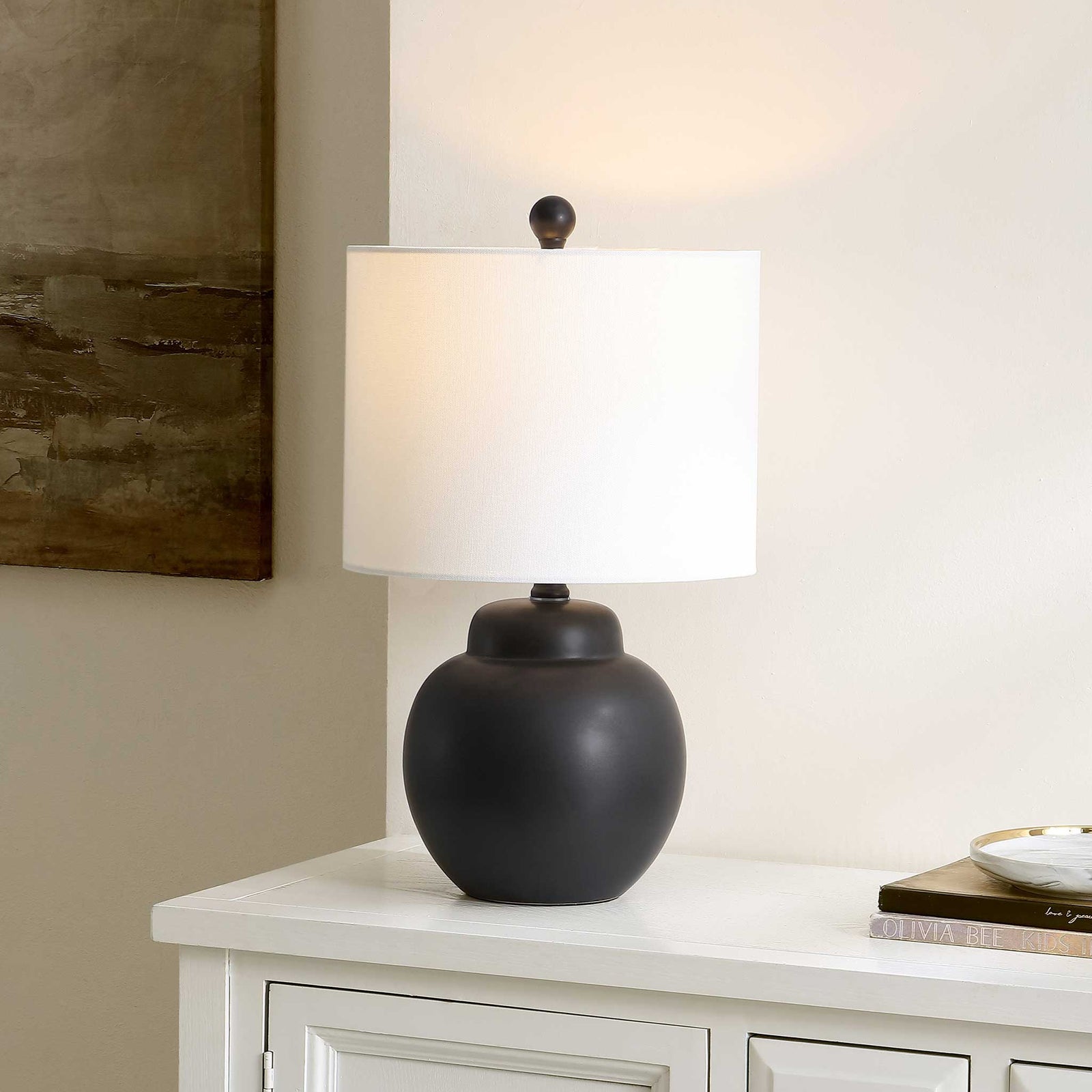 Illuminated black ceramic lamp casting a warm glow through its white drum shade || TBL4513A || front_angle_on_room