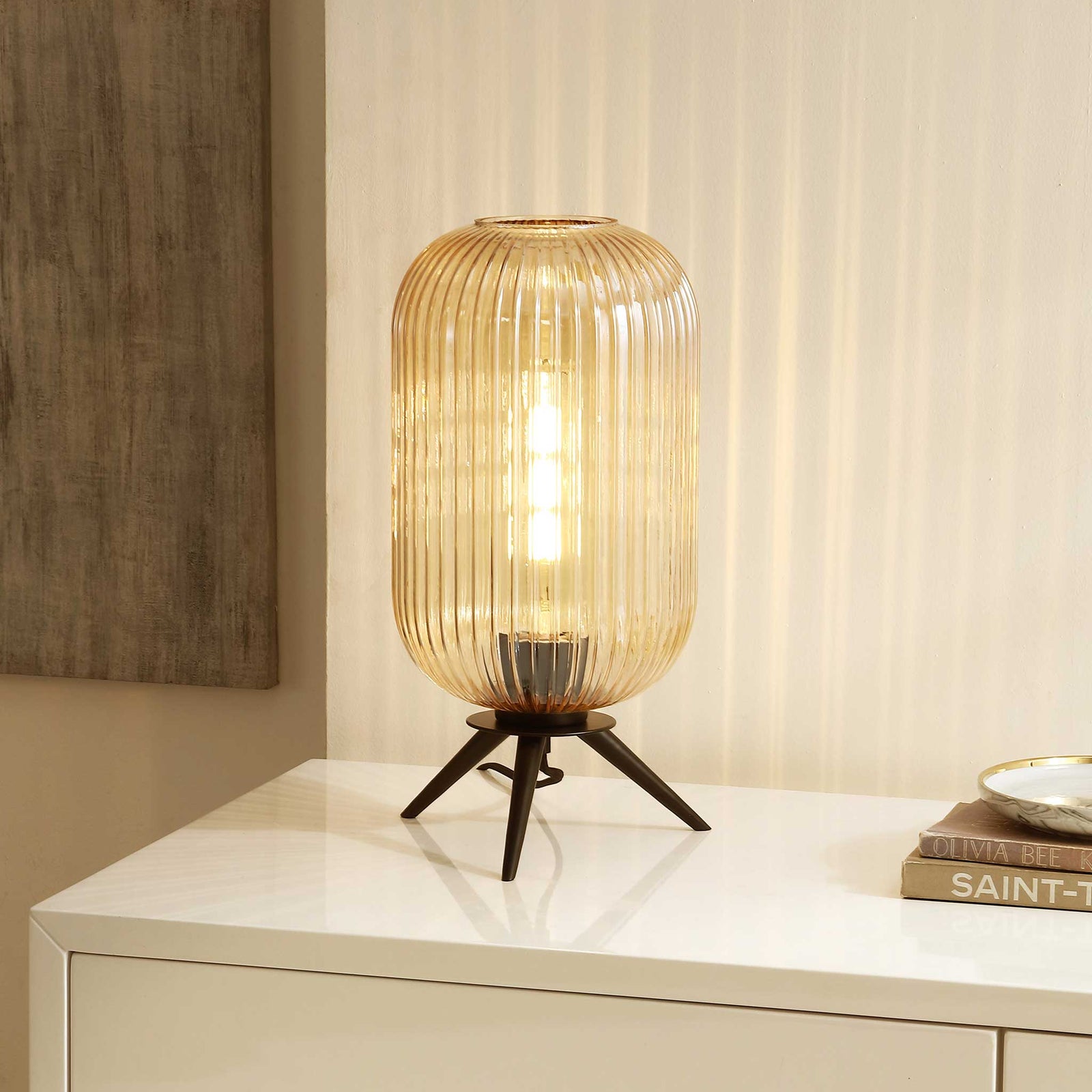 Soft, inviting light shines through ribbed amber glass on a stable black three-leg base. || TBL4528A || front_angle_on_room