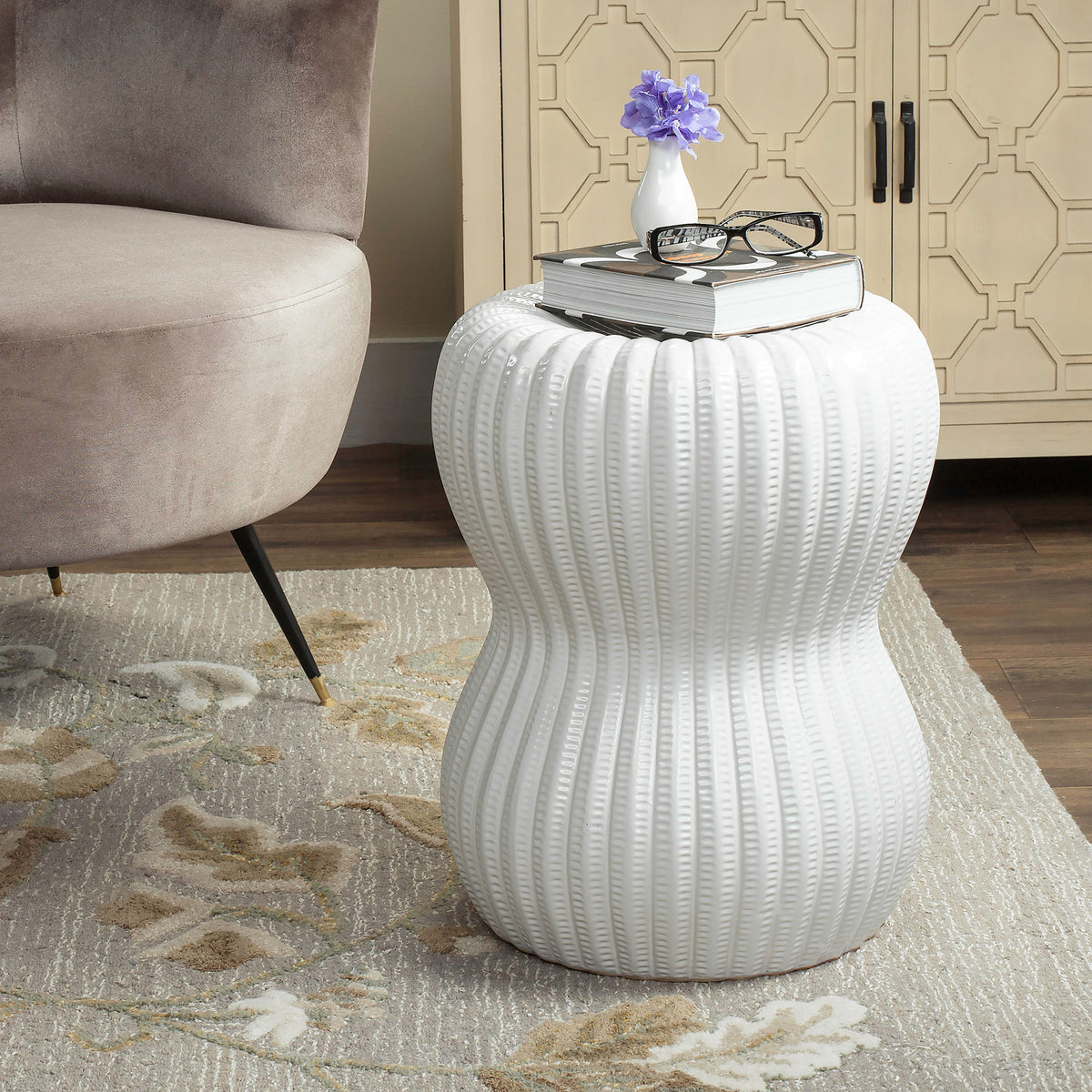 A white ceramic garden stool with hourglass curves holds books and blooms beside a chair. || ACS4518A || front_angle_room