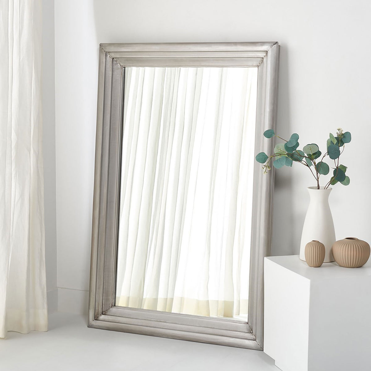A large silver-framed mirror leans against a bright wall, reflecting soft white curtains. || CMI2001B || front_room