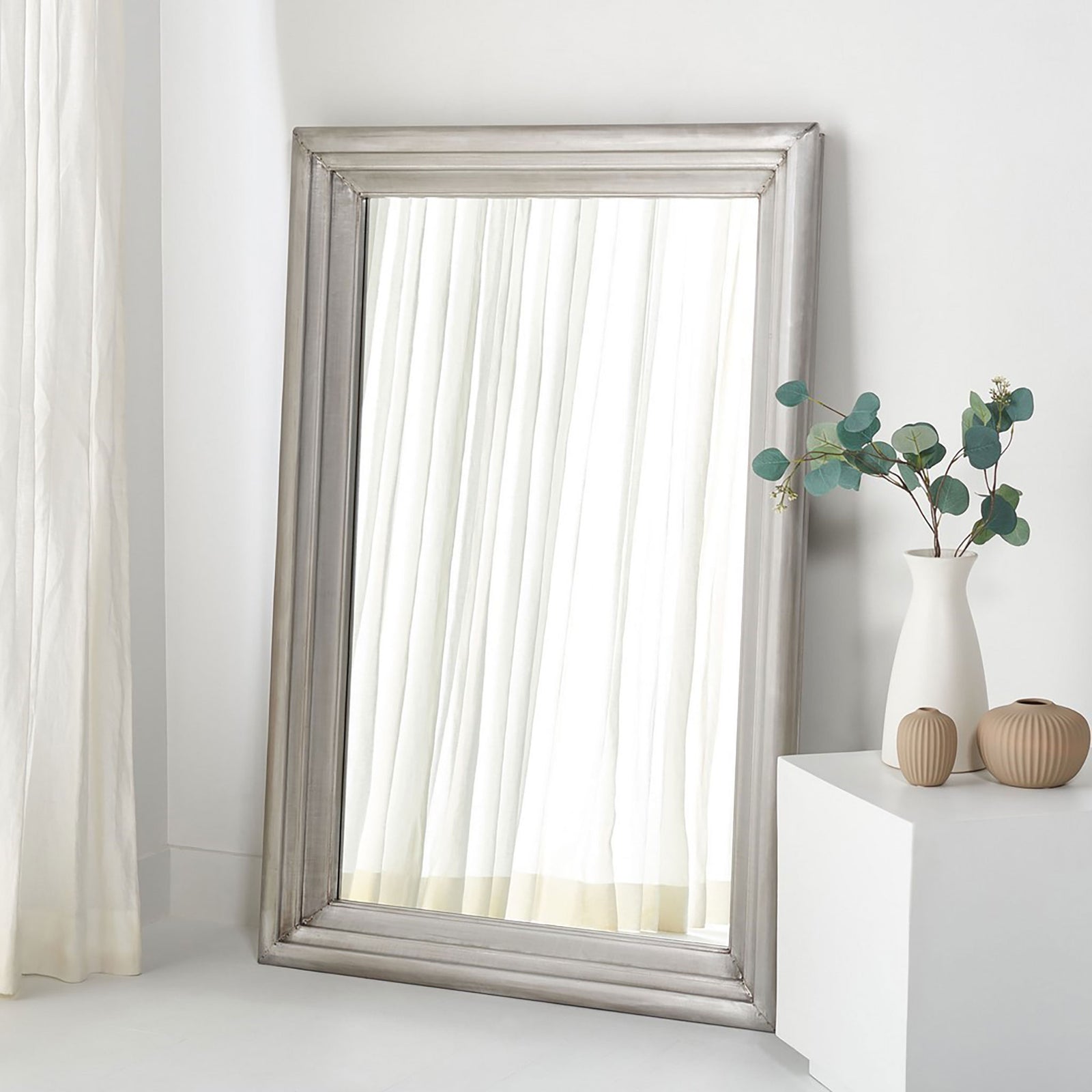 A large silver-framed mirror leans against a bright wall, reflecting soft white curtains. || CMI2001B || front_room