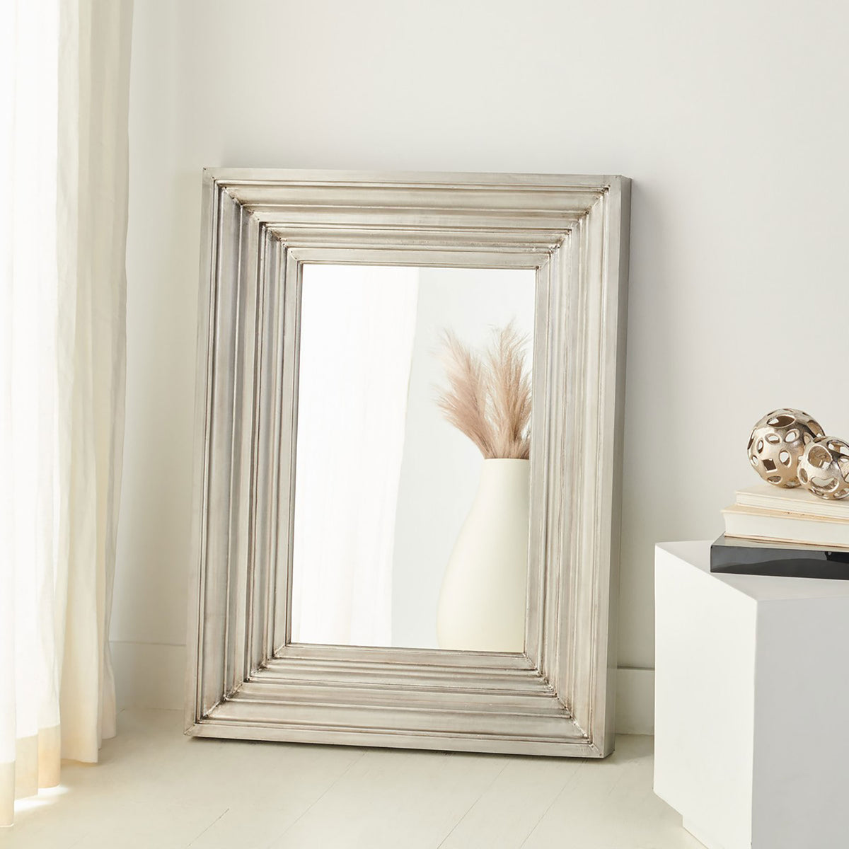 Wide silver frame adds sculptural detail as the mirror reflects a vase of dried stems. || CMI2004B || front_room