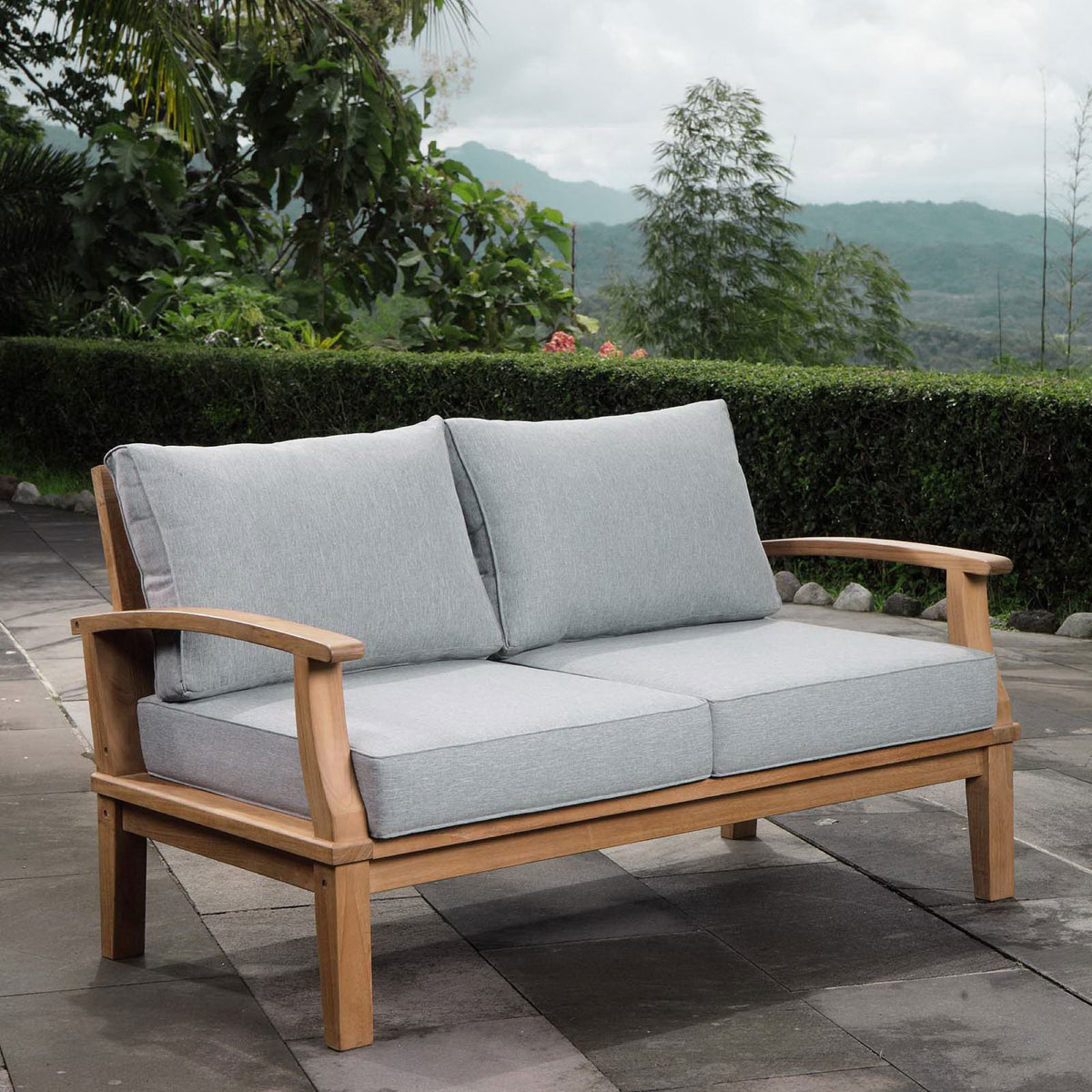 Teak loveseat with gray cushions styled on a stone patio amid lush outdoor greenery || EEI-1144-NAT-GRY-SET || front_angle_room