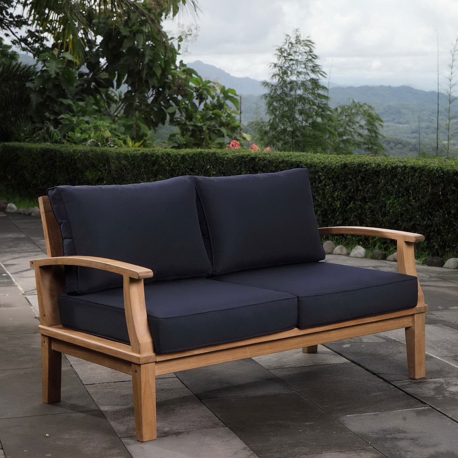 Teak loveseat with navy cushions set on a terrace overlooking lush hills and gardens || EEI-1144-NAT-NAV-SET || front_angle_room