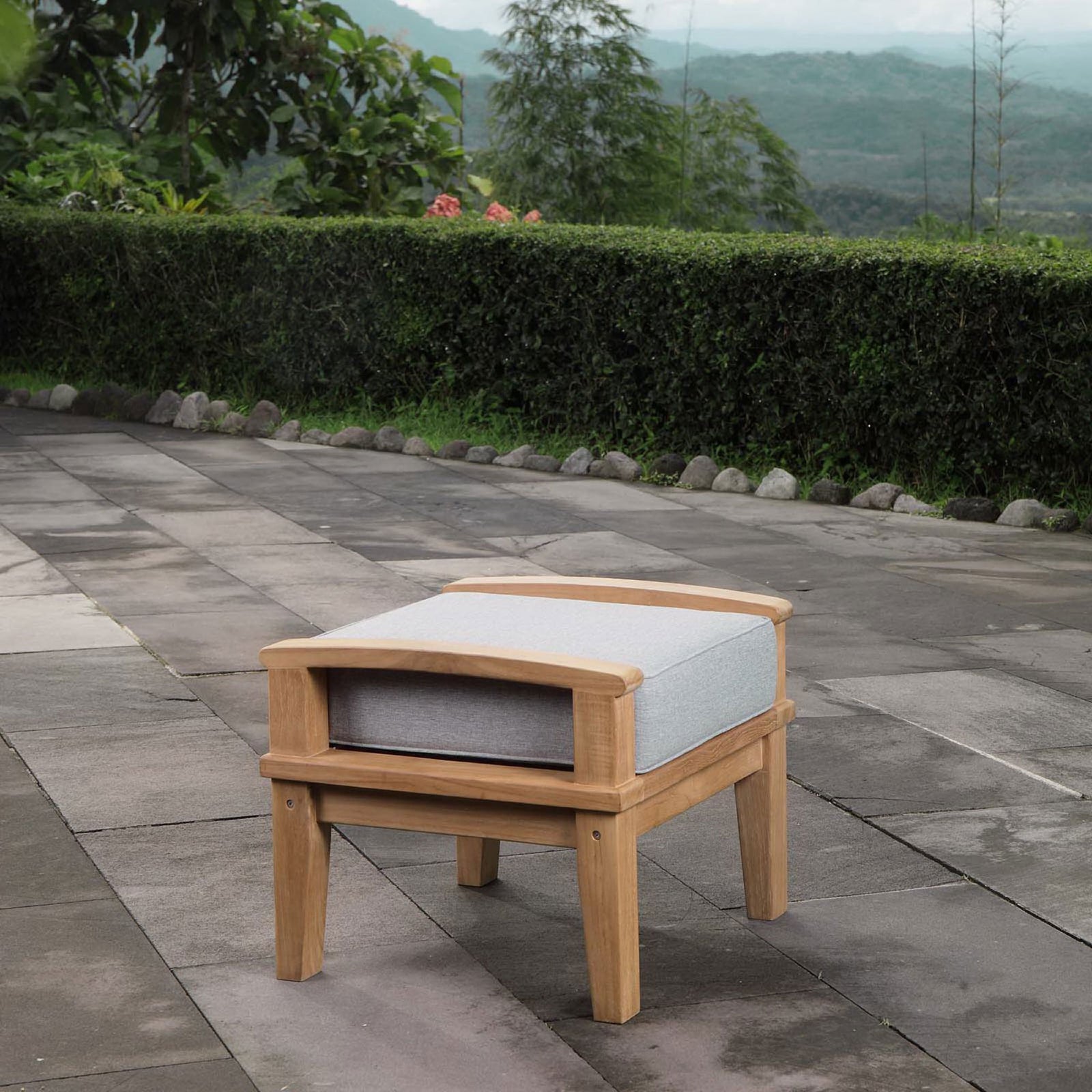 Teak ottoman with gray cushion styled on a stone patio surrounded by lush greenery. || EEI-1152-NAT-GRY-SET || front_angle_room