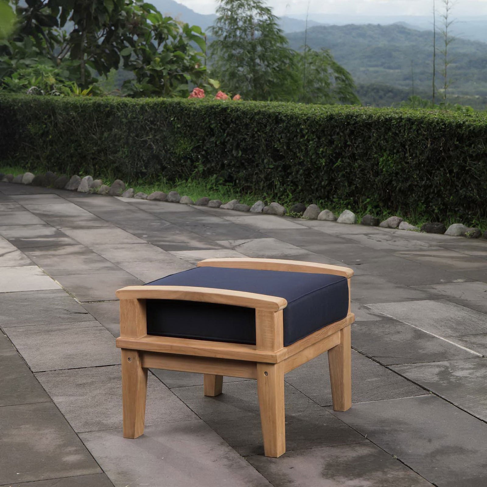Teak ottoman with navy cushion styled on a stone patio overlooking lush greenery. || EEI-1152-NAT-NAV-SET || front_angle_room