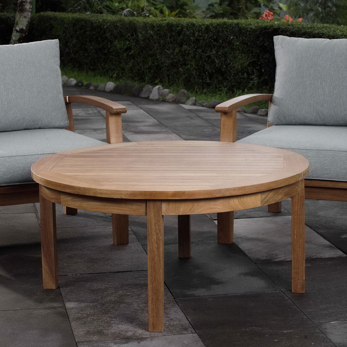 Round teak coffee table arranged on a patio between lounge chairs for outdoor relaxation. || EEI-1153-NAT || front_angle_room