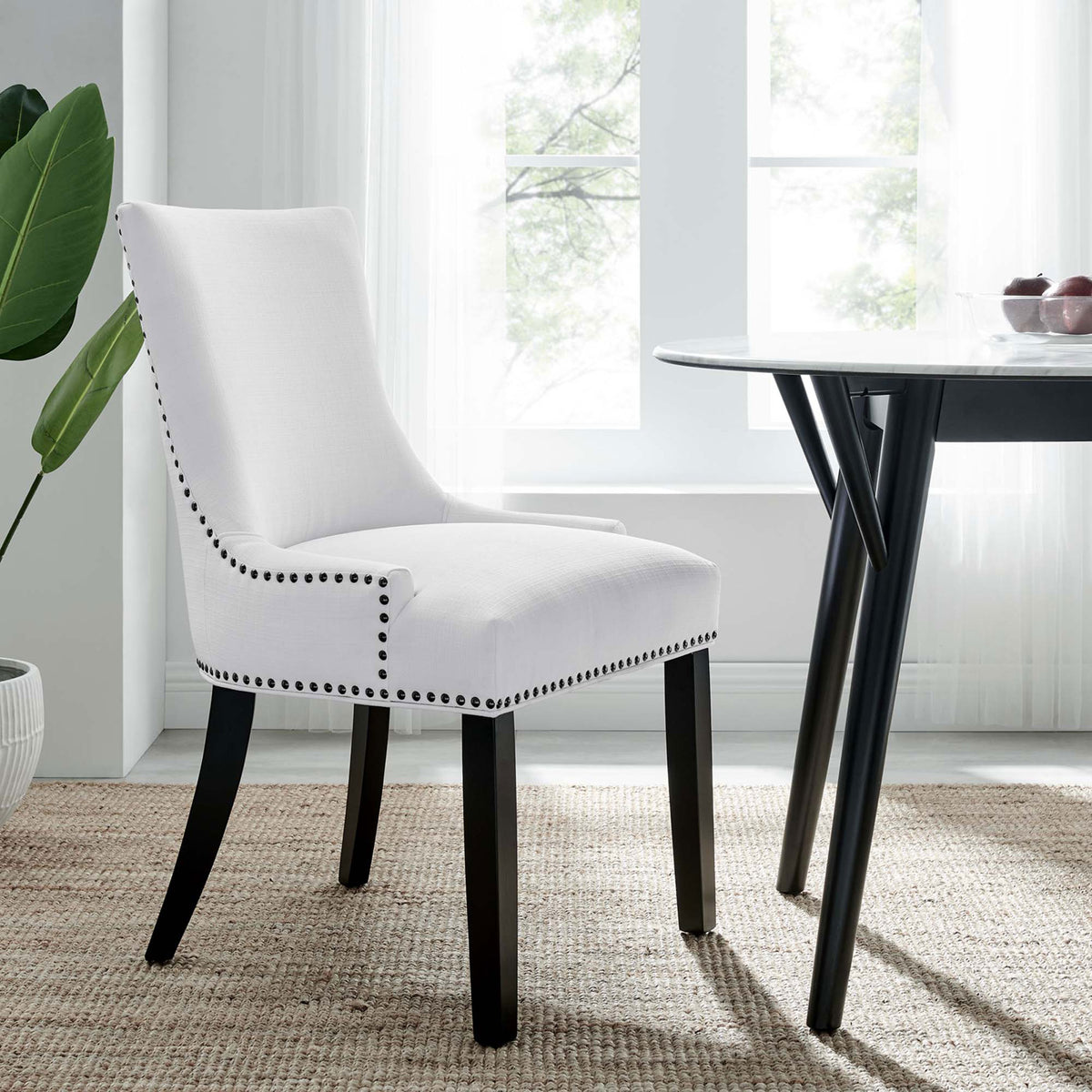 White upholstered dining chair with nailhead trim styled beside a round dining table in a bright airy room || EEI-2229-WHI || front_angle_room