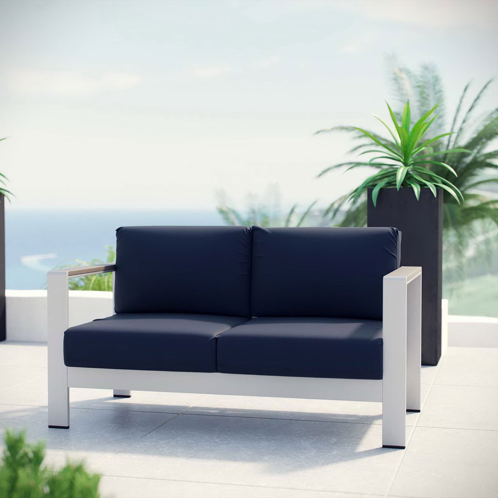 Navy cushioned aluminum loveseat arranged on a bright patio with lush planters and ocean view || EEI-2267-SLV-NAV || front_room