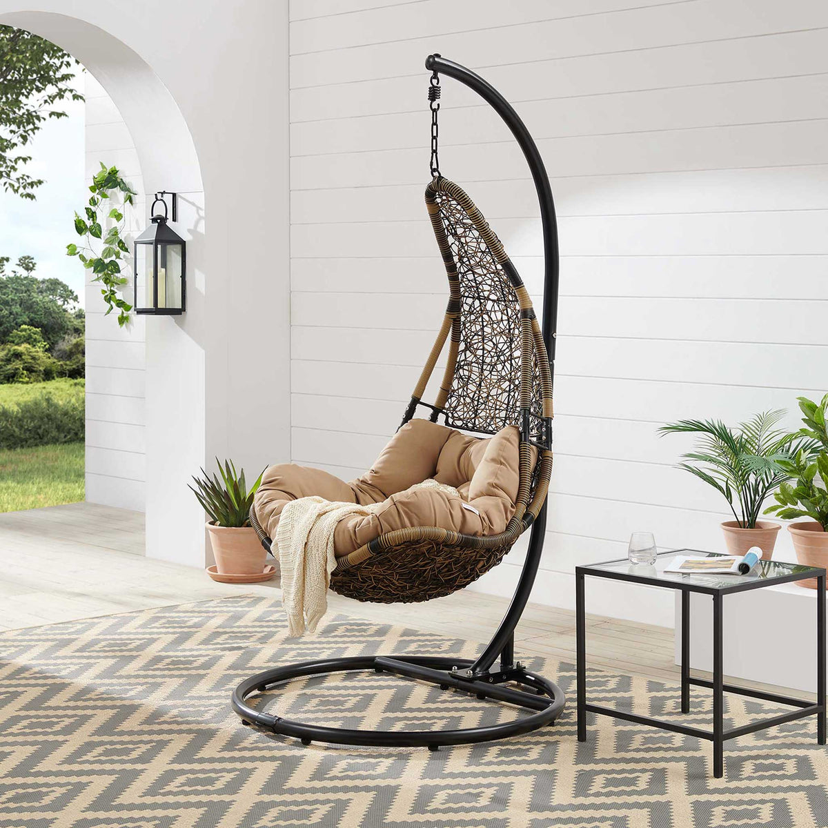 Brown wicker patio swing chair with mocha cushion styled on a light filled covered patio.