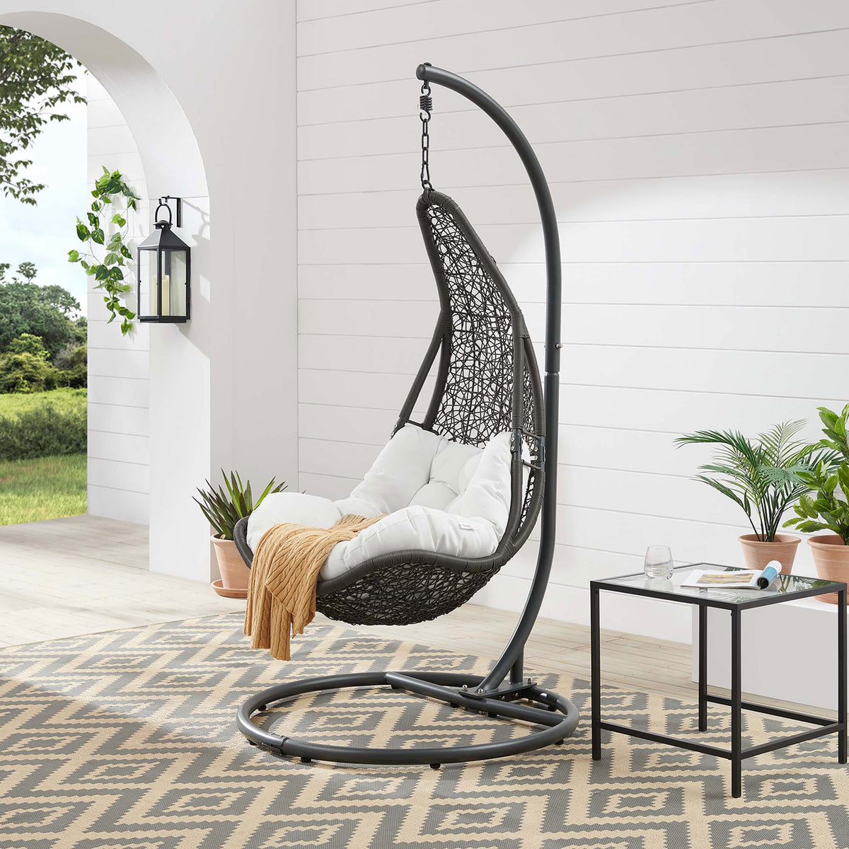 Gray wicker patio swing chair with white cushion styled on a covered terrace with greenery. || EEI-2276-GRY-WHI-SET || front_angle_room
