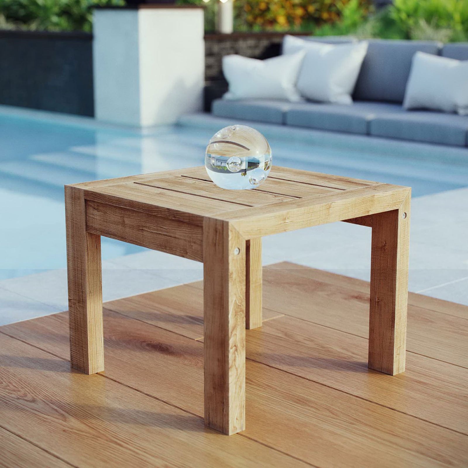 Teak outdoor side table set beside a modern poolside lounge with decorative glass orb accent. || EEI-2709-NAT || front_angle_room