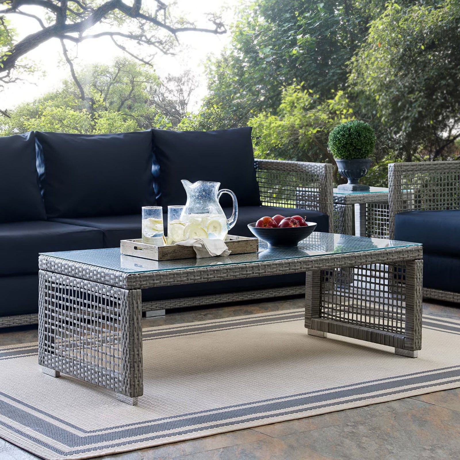 Gray wicker outdoor coffee table with glass top styled on a patio with coordinating seating and fresh refreshments || EEI-2919-GRY || front_angle_room