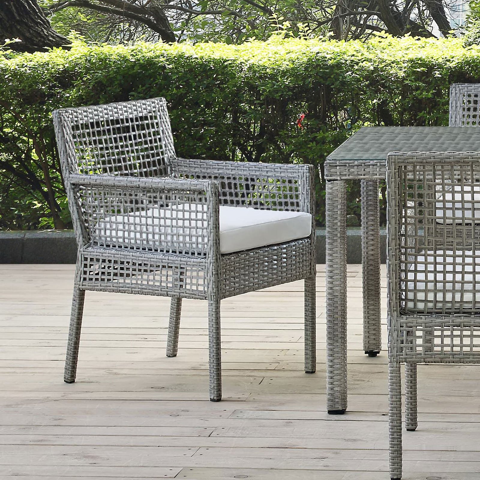 Gray wicker outdoor dining armchair with white cushion styled on a wood deck beside a matching dining table || EEI-2920-GRY-WHI || front_angle_room