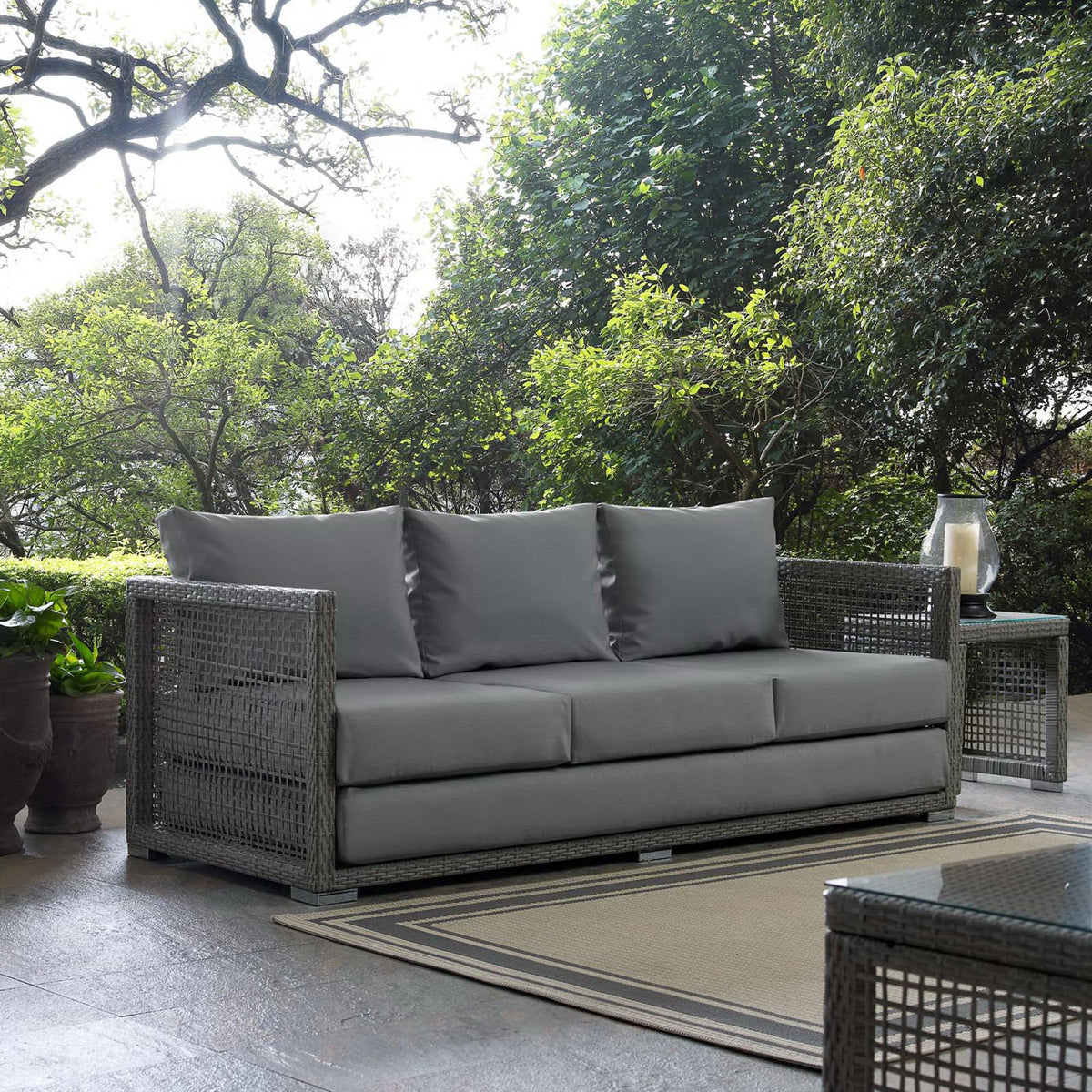 Gray wicker outdoor sofa with plush gray cushions styled on a patio beside matching tables and greenery || EEI-2923-GRY-GRY || front_angle_room
