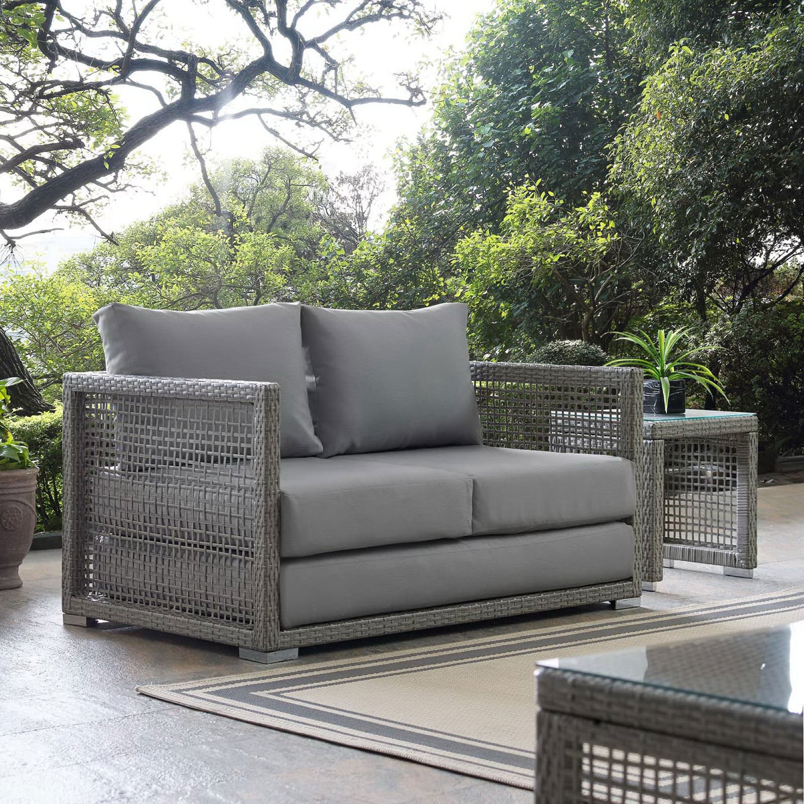 Gray wicker outdoor loveseat styled on patio with matching tables and greenery || EEI-2924-GRY-GRY || front_angle_room