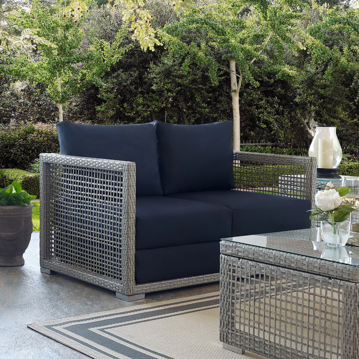 Gray wicker loveseat with navy cushions styled on a lush outdoor patio || EEI-2924-GRY-NAV || front_angle_room