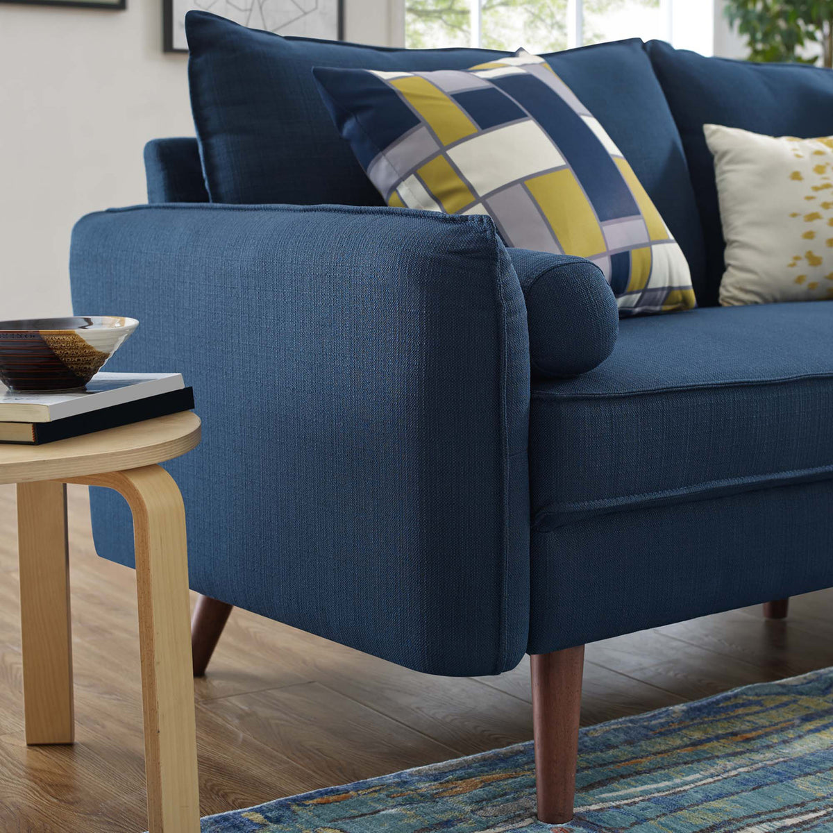 Textured blue upholstery wraps the broad arm beside a soft bolster pillow and a tapered wood leg.