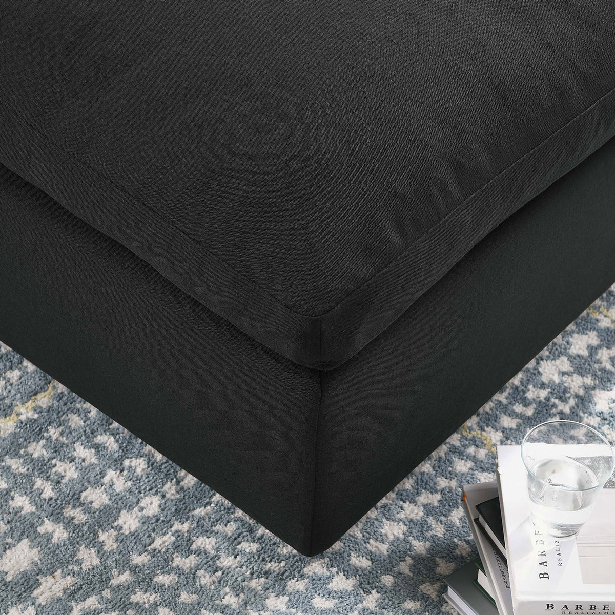 Corner of the black ottoman styled on a patterned rug beside books and a glass of water. || EEI-3318-BLK || detail_room