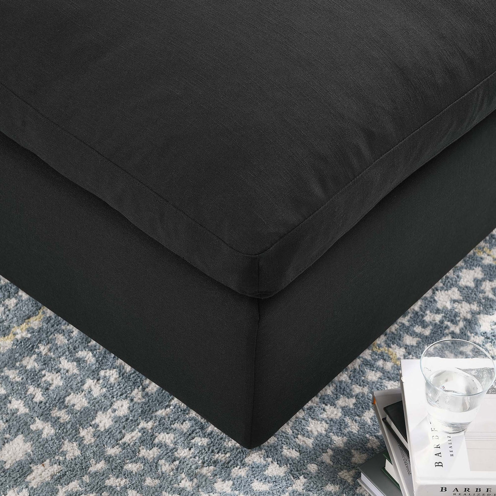Corner of the black ottoman styled on a patterned rug beside books and a glass of water. || EEI-3318-BLK || detail_room