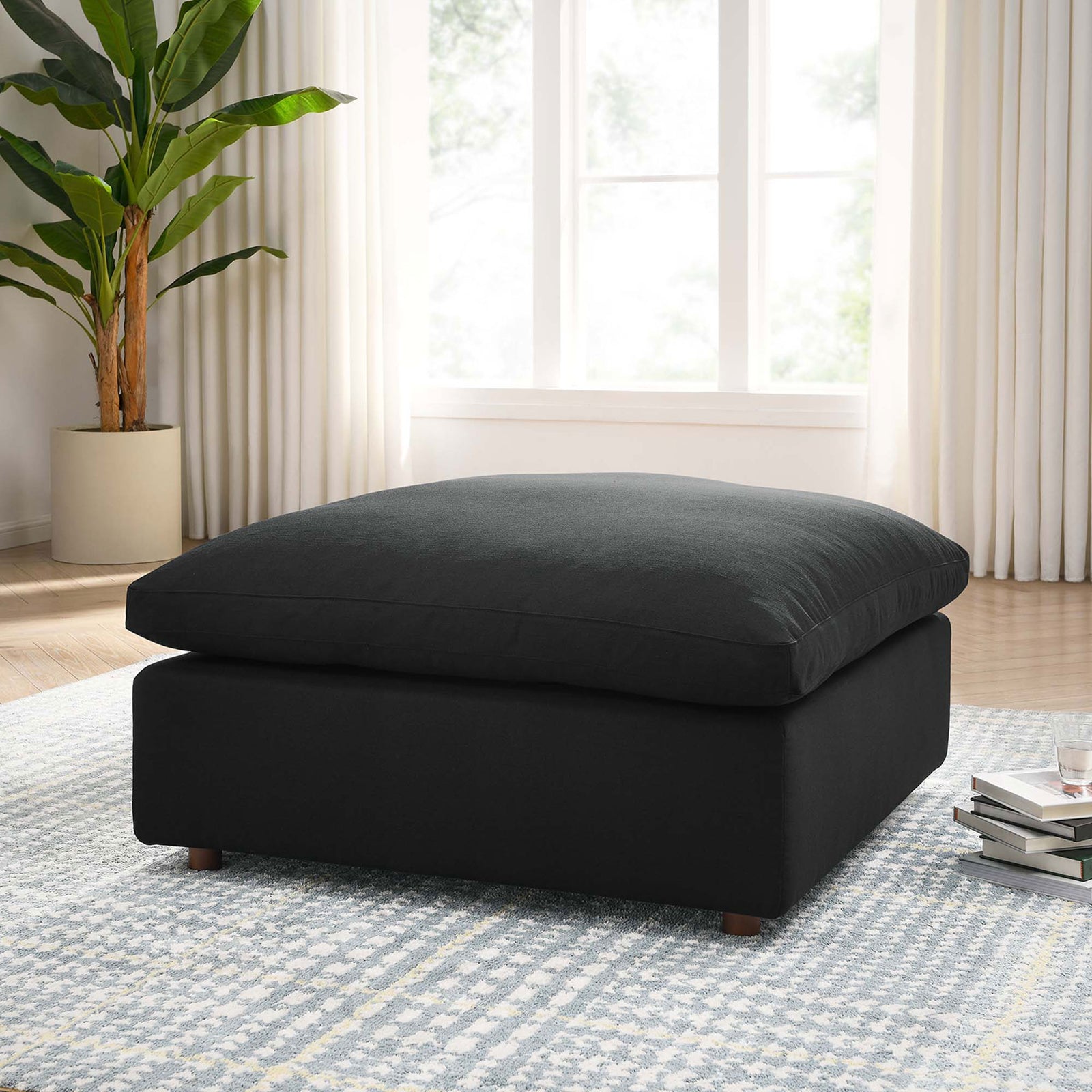 Black overstuffed ottoman styled in a bright living room, layered over a patterned rug with modern decor. || EEI-3318-BLK || front_angle_room