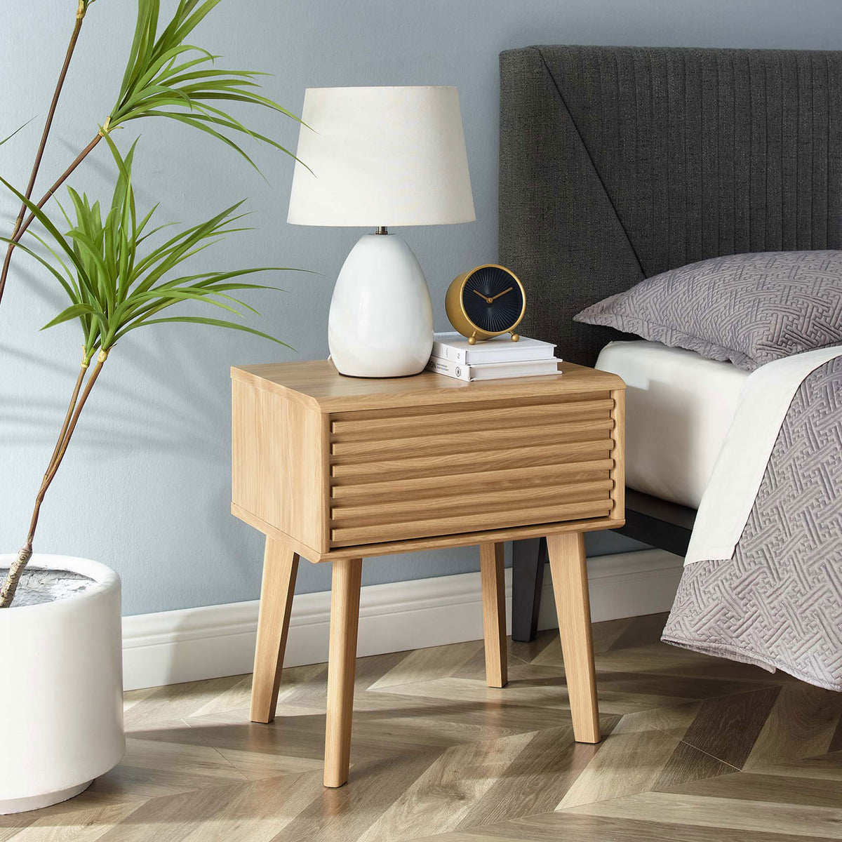 Oak nightstand styled next to bed with lamp, clock, and books on the tabletop || EEI-3345-OAK || front_angle_room